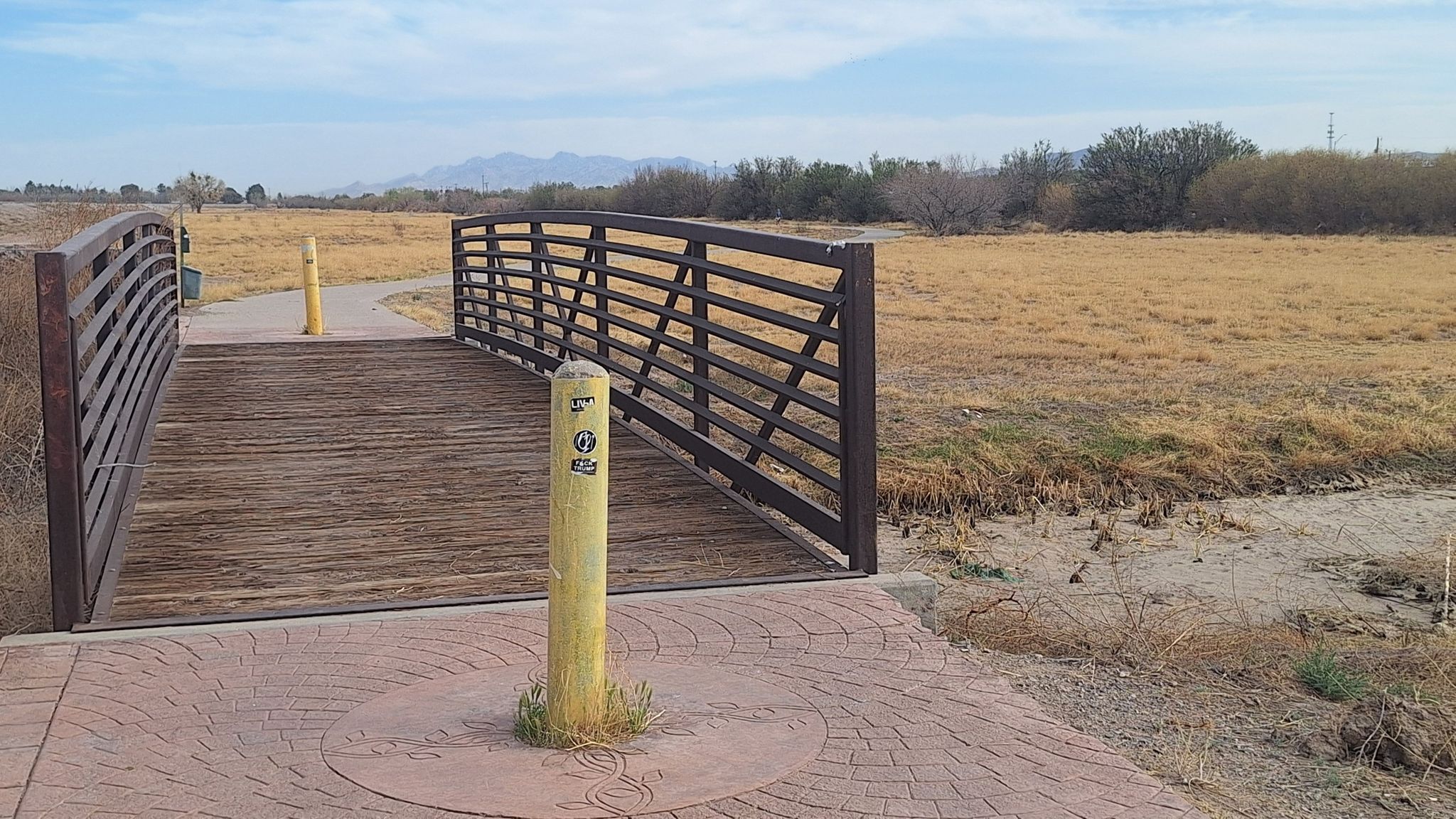 A wooden and metal pedestrian bridge crosses a dry ditch in a sunny field. - River Park Trail