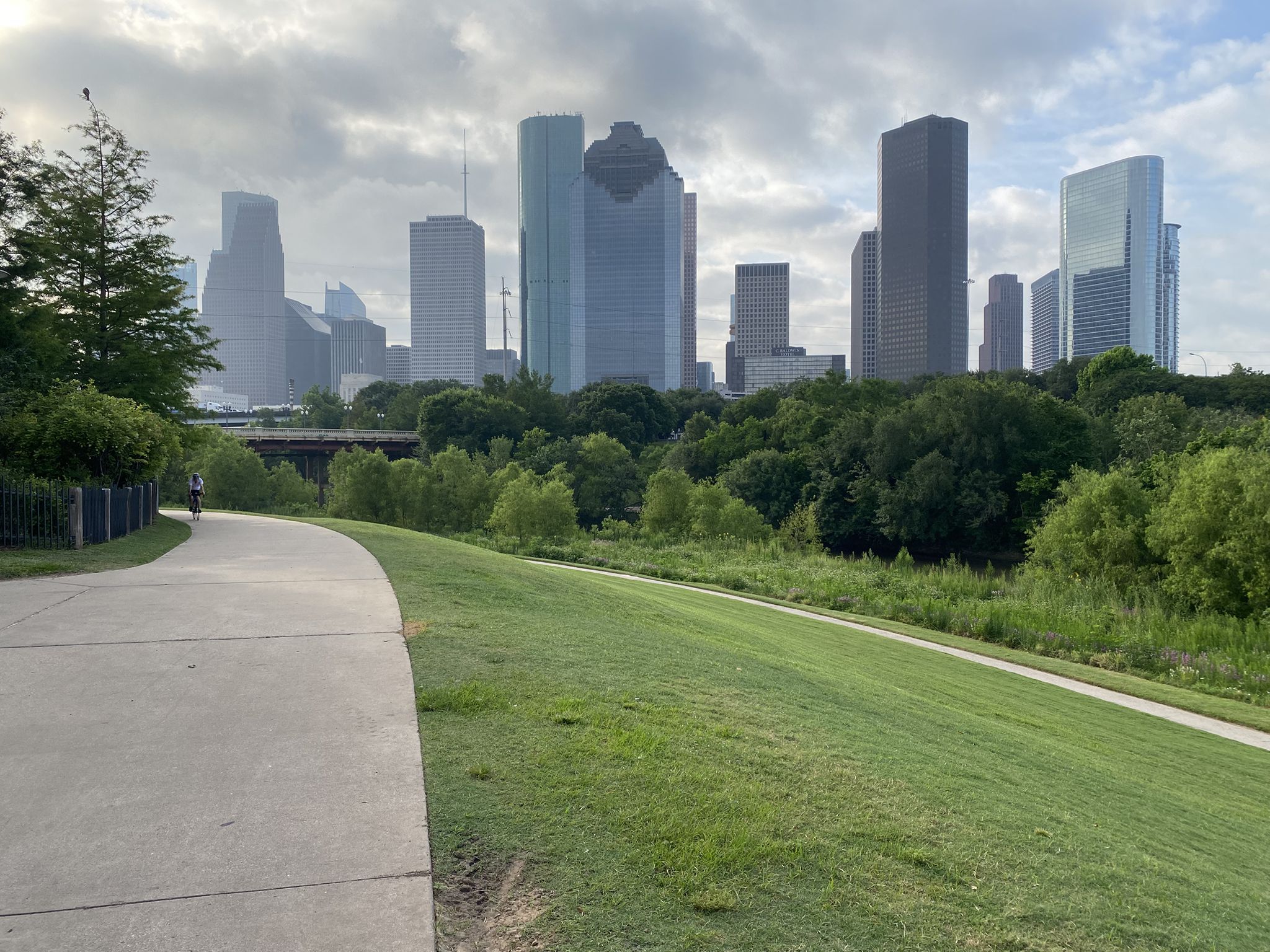 Paved path curves beside a grassy hill with a distant city skyline. - Buffalo Bayou Park – The Cistern at Buffalo Bayou Park loop from Theater District (eastbound)