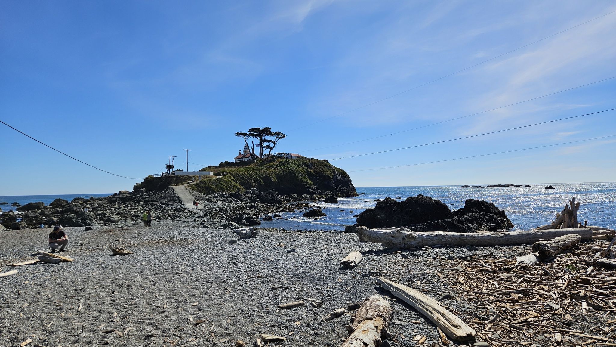 Sunny rocky beach with driftwood, ocean, and a distant tree-covered island. - Battery Point Lighthouse
