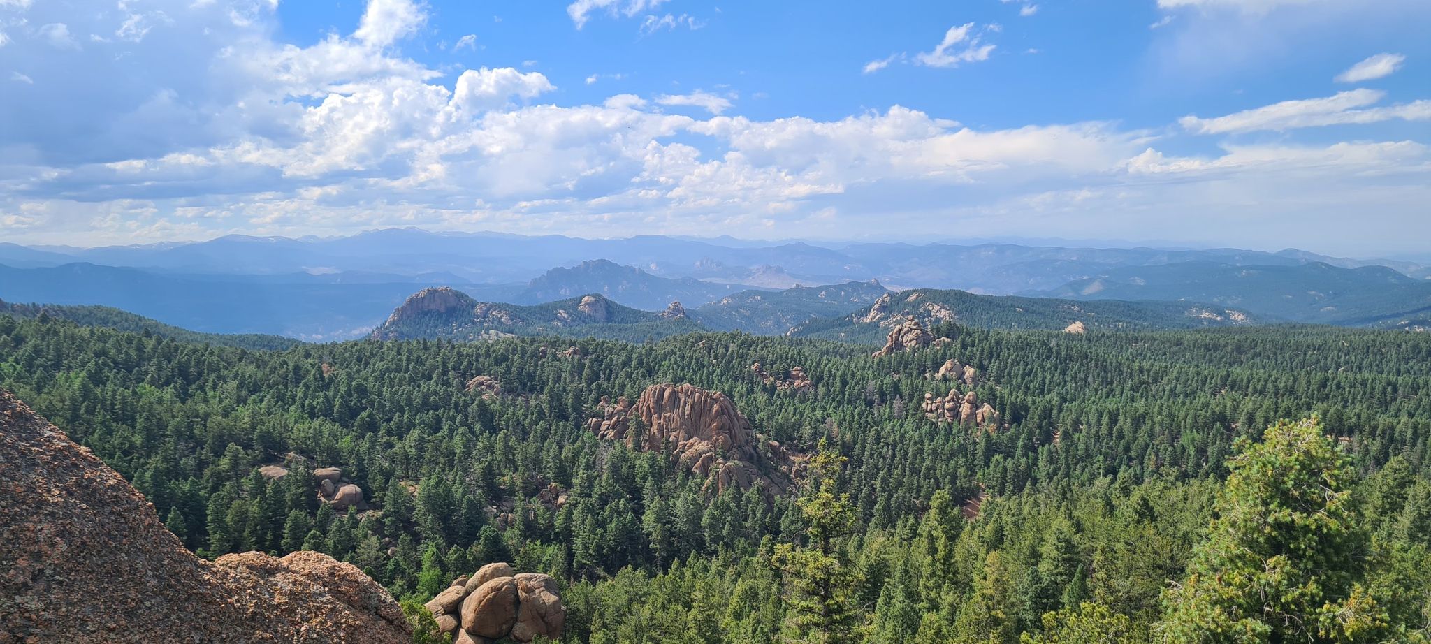 Vast evergreen forest and rocky mountains under a bright, partly cloudy sky. - Devil's Head Trail