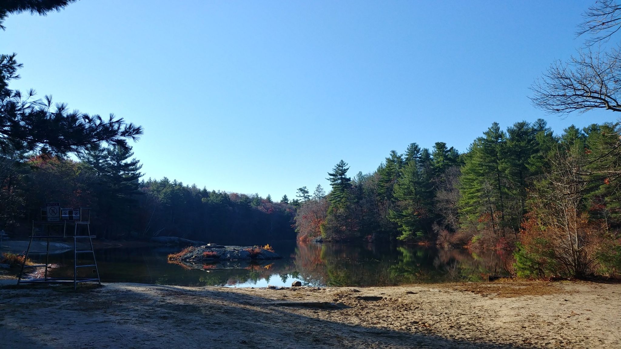 Sunny view of a calm lake surrounded by dense forest and a sandy shore. - Ridge and Saugus River Trail Loop