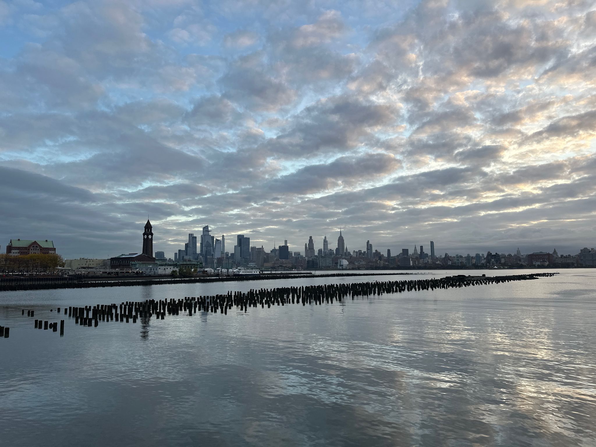 Distant city skyline across water with a line of wooden posts. - Liberty State Park – LeFrak Point Lighthouse loop from Hoboken Terminal