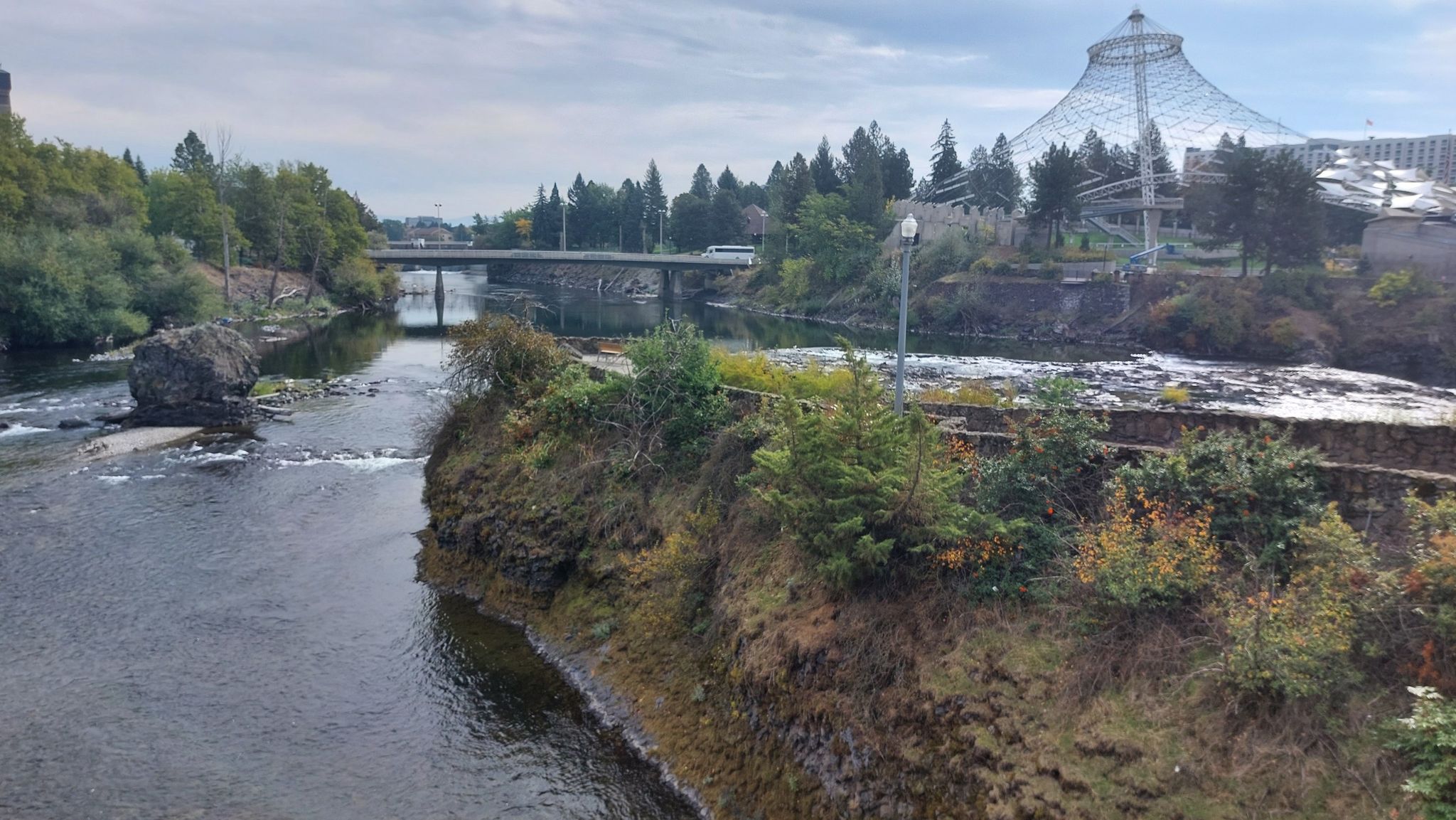 Overcast view of a river flowing under a bridge, with a large glass structure. - Spokane Falls loop