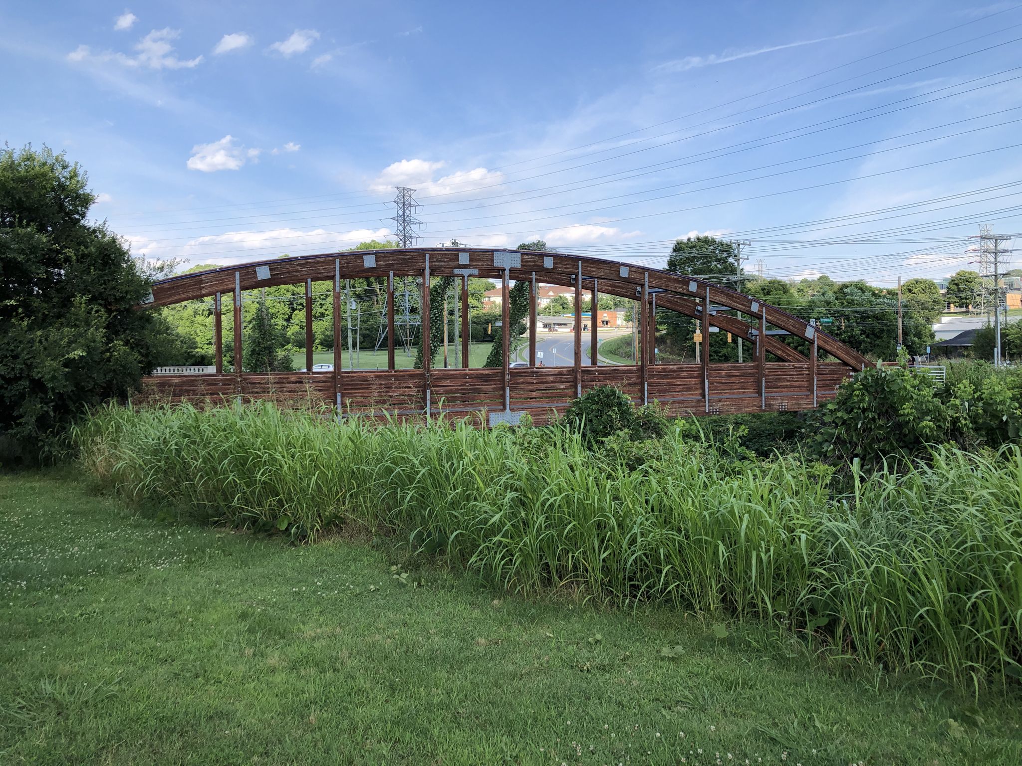 A modern wooden pedestrian bridge spans over a grassy area on a sunny day. - Long Branch Trail – Molly Leight's Crossing loop from Old Salem