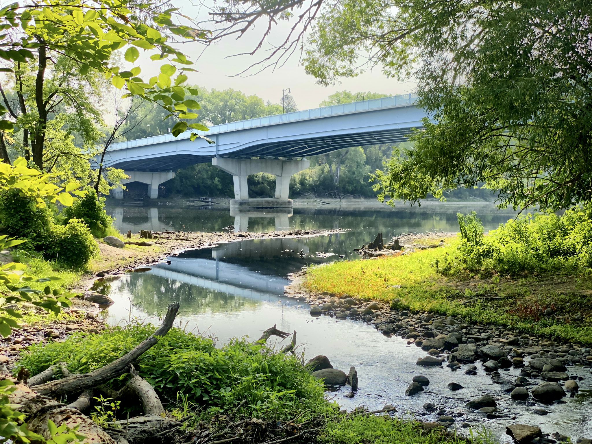 A large bridge spans a calm river with green banks under bright, dappled sunlight. - North Mississippi Park Loop