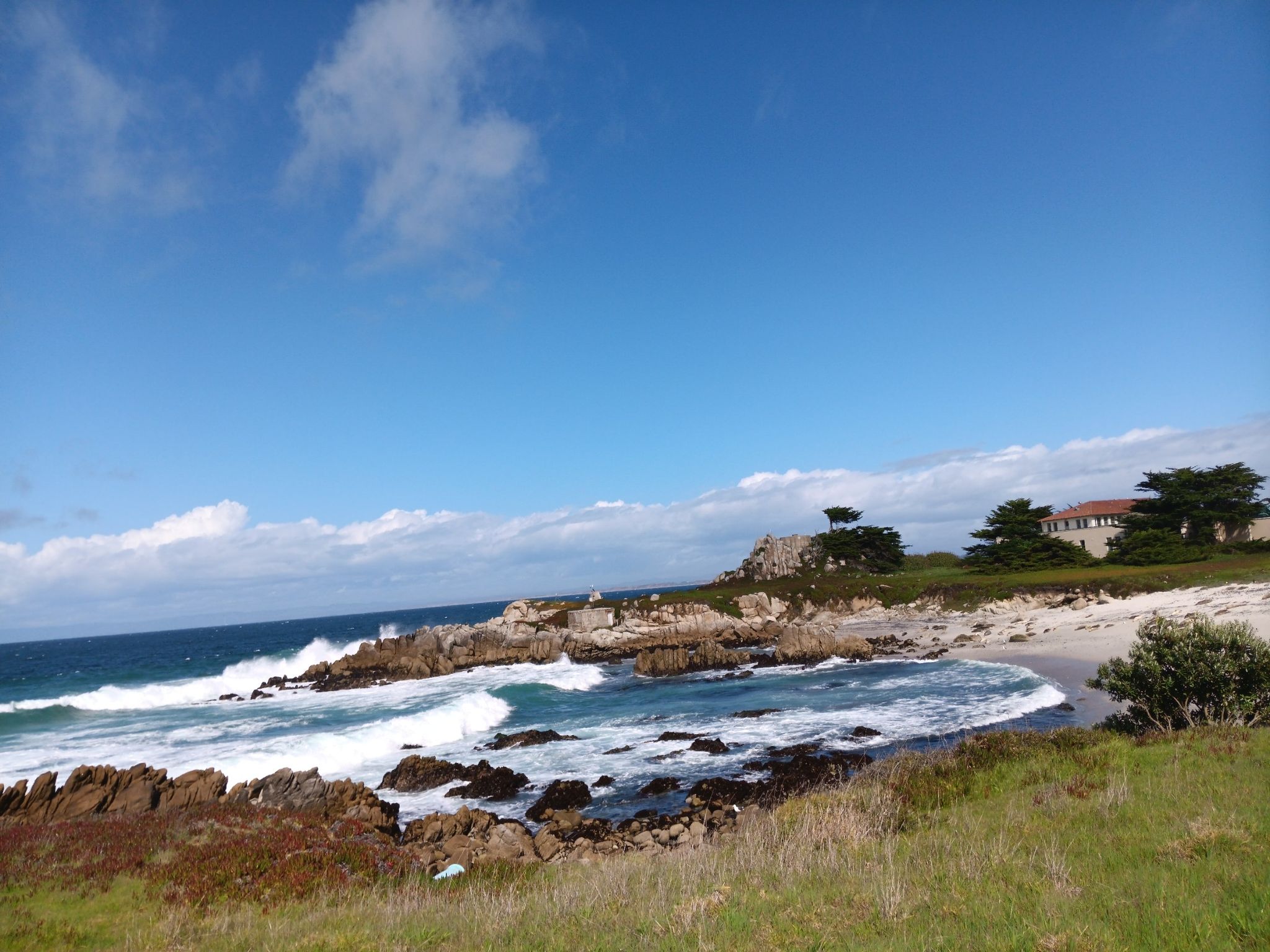 Sunny day with blue sky, crashing waves on a rocky shore, and distant buildings. - Monterey Bay Coastal Recreation Trail