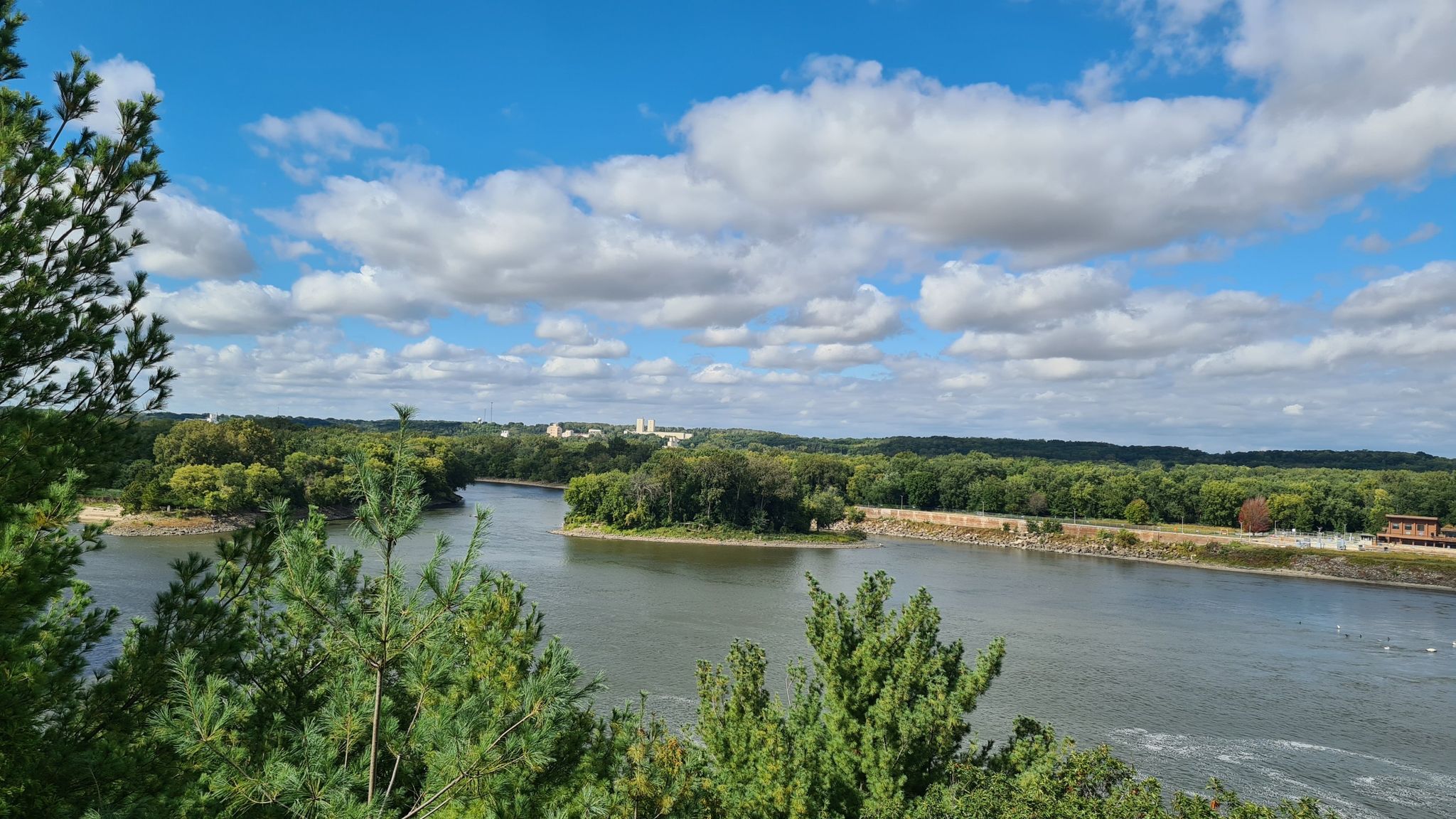 Wide river view with forested banks, distant buildings, and a cloudy blue sky. - River Trail, Bluff Trail, and Illinois Canyon Loop