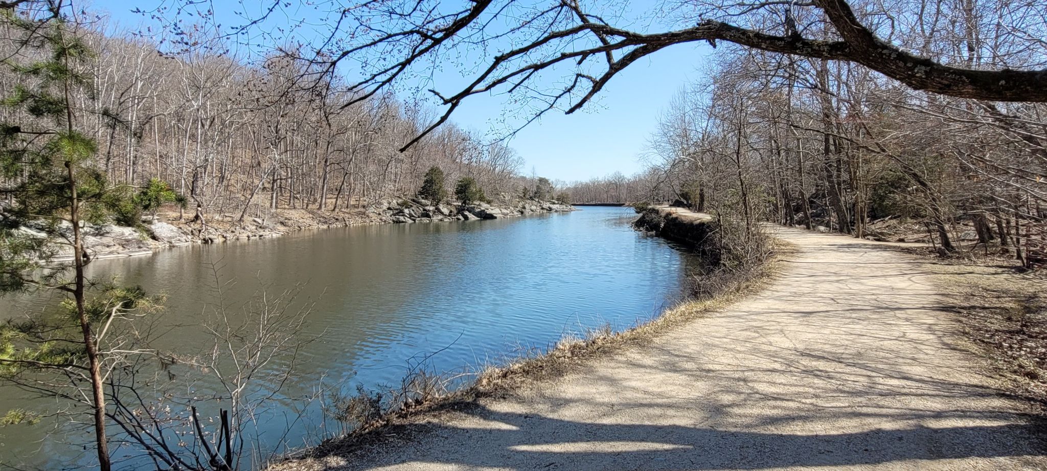 Calm river next to a dirt path, surrounded by bare trees under a blue sky. - Billy Goat A Trail Entrance loop from Chesapeake and Ohio Canal National Historical Park