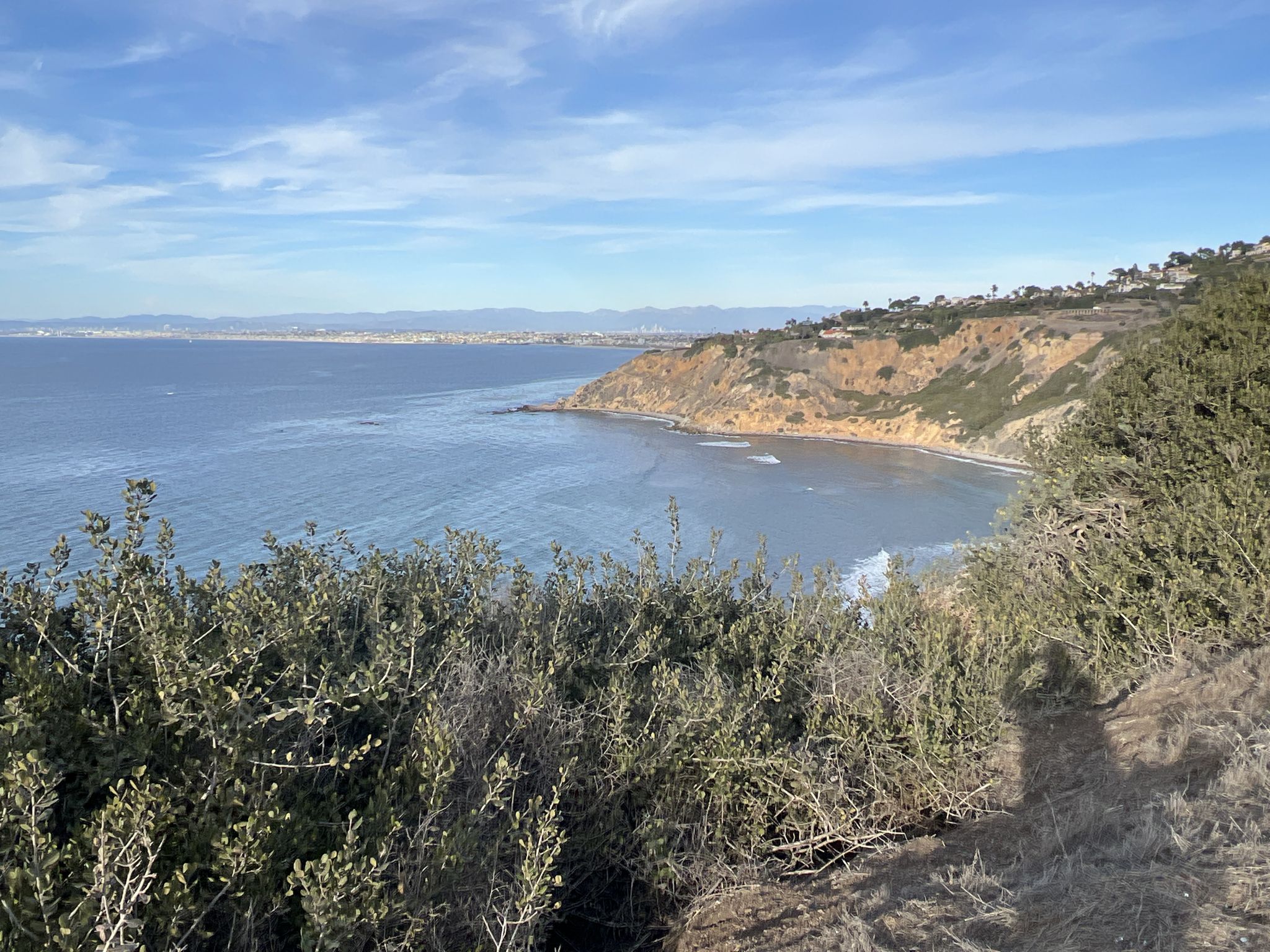 Sunny coastal view with blue ocean, rocky cliffs, green bushes, and a person's shadow. - Malaga Lunada Vista Loop
