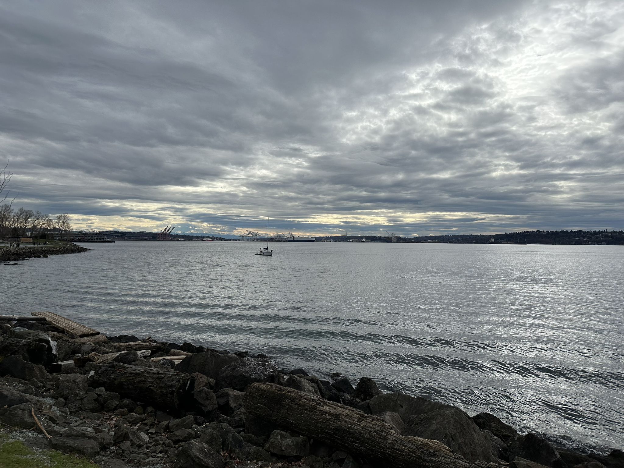 Wide bay with small boat, rocky shore, and distant land under cloudy sky. - Myrtle Edwards Park – Elliott Bay Trail loop from Climate Pledge Arena