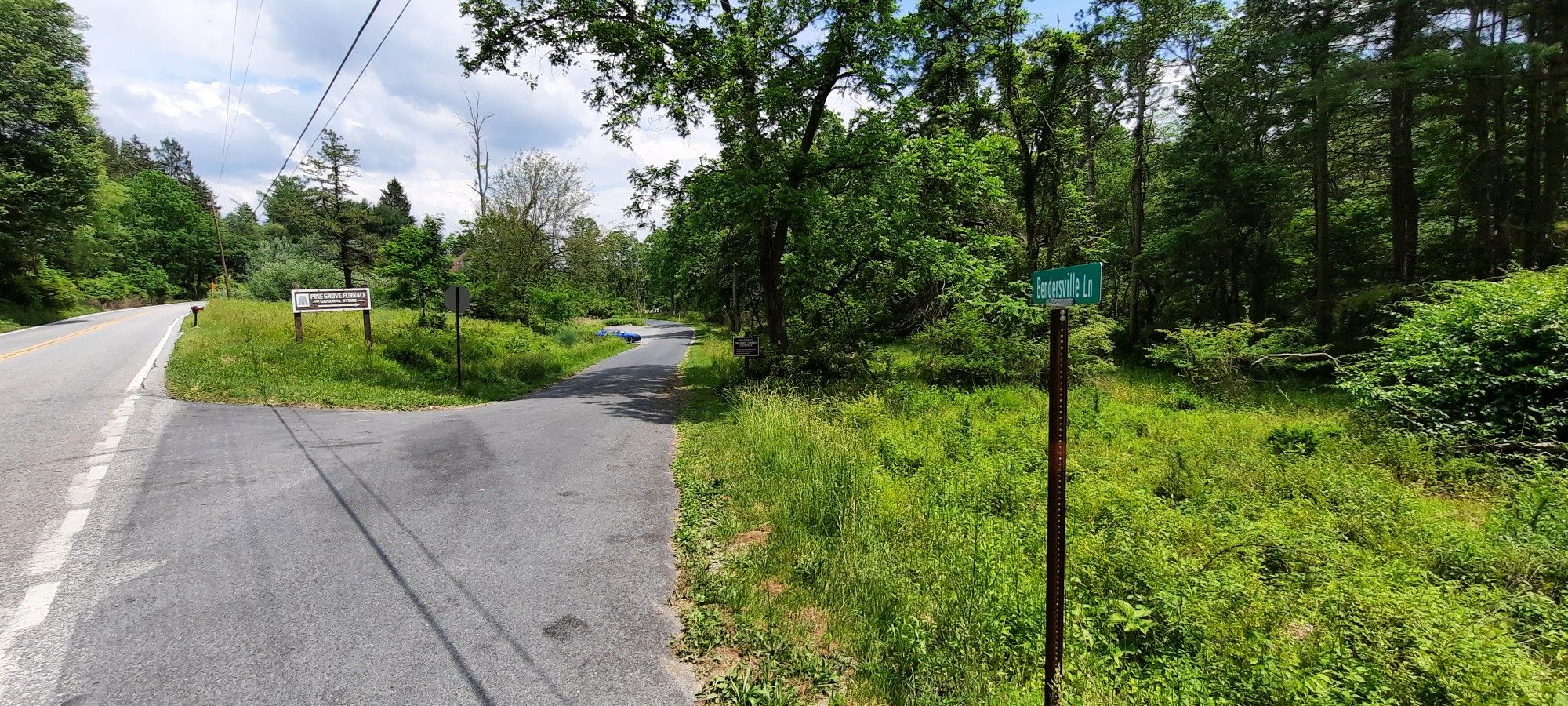 A paved road intersection with a dirt path, surrounded by green grass and trees. - Appalachian Trail Museum – Iron Furnace loop from Laurel Lake