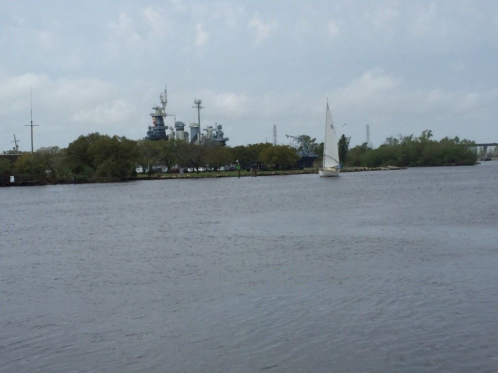 Distant warship and a sailboat on a wide river under a cloudy sky. - Wilmington Historic District – View of USS North Carolina loop from Wilmington