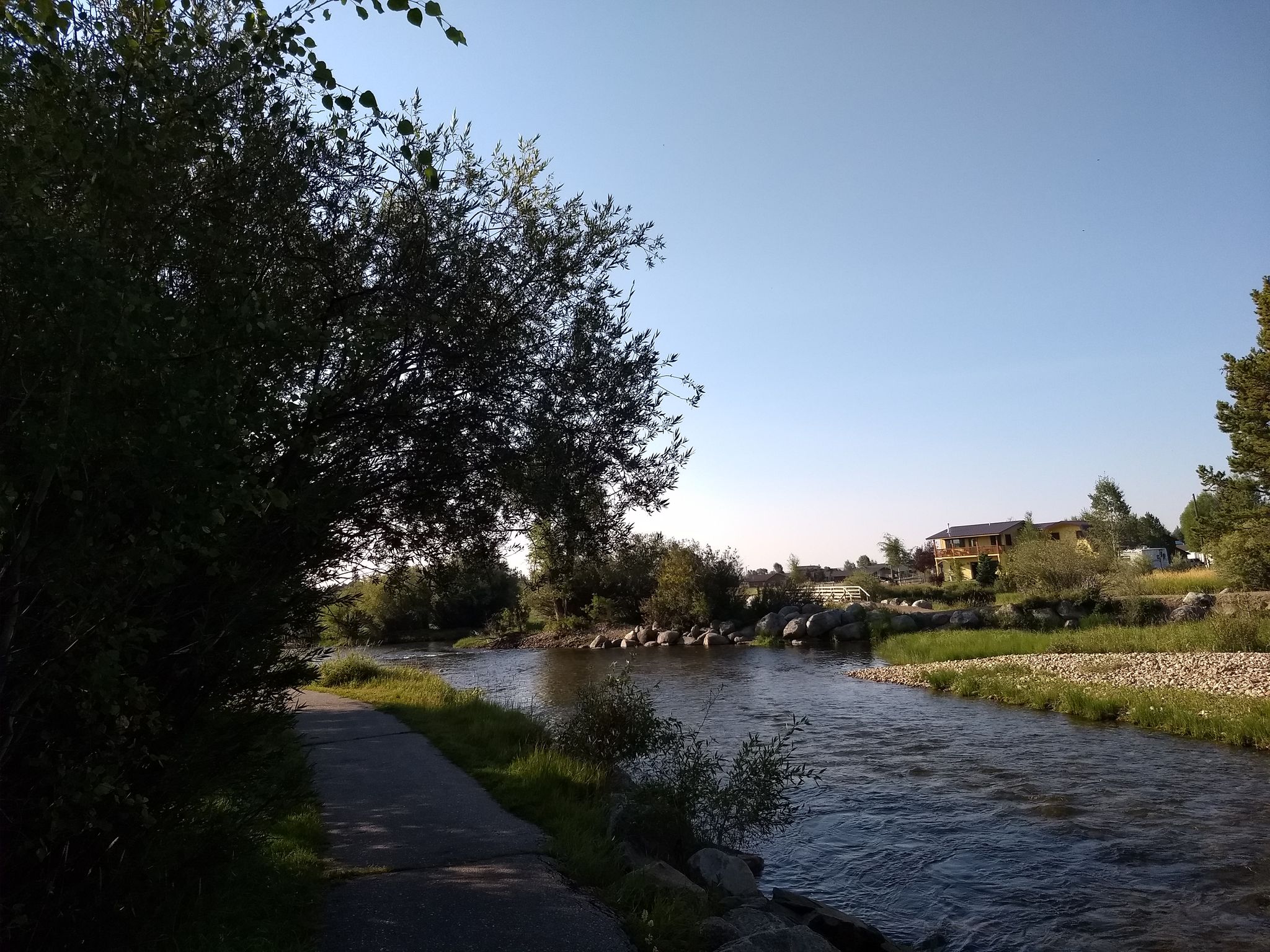 A paved path runs alongside a river with trees and distant buildings. - Boyd Skinner Trail