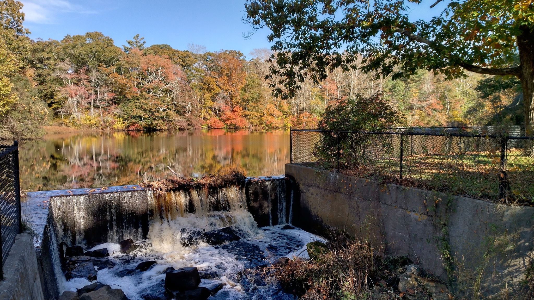Waterfall over a concrete dam, with a lake reflecting autumn trees under a sunny sky. - View of Westport Point – East Horseneck Beach loop from Smith Farm