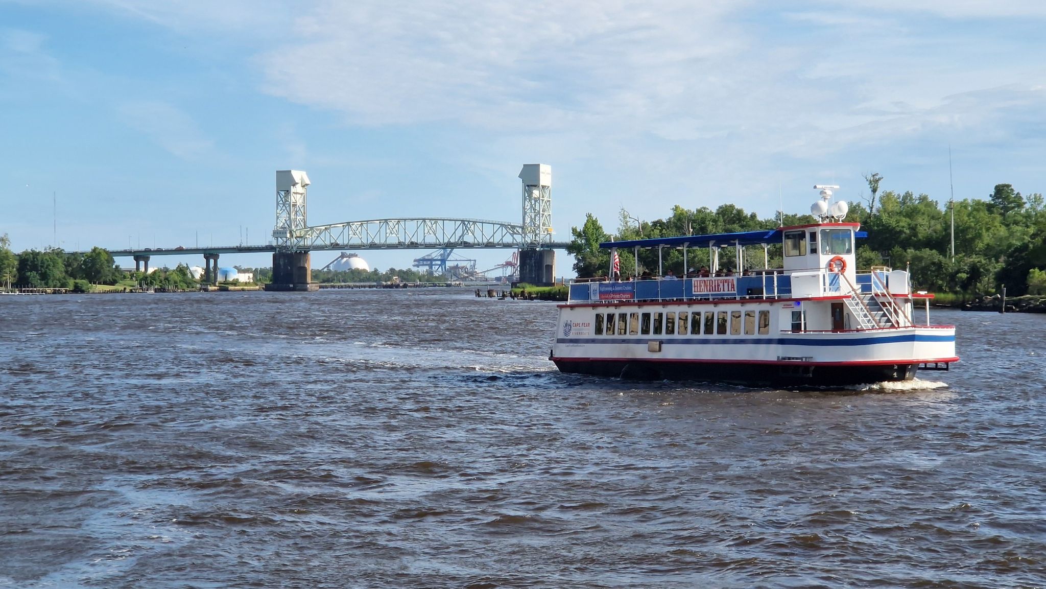 Passenger boat on a river with a large metal bridge in the background. - View of USS North Carolina – Wilmington Historic District loop from Wilmington