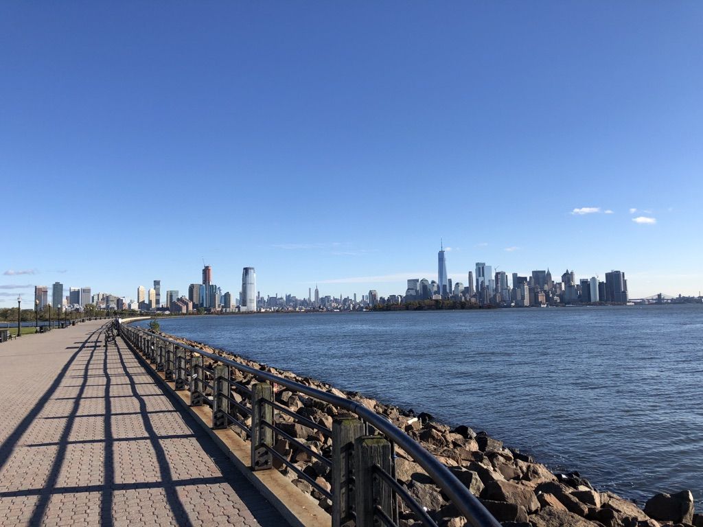 Paved waterfront path with city skyline across the water under blue sky. - Richard J. Sullivan Natural Area Loop