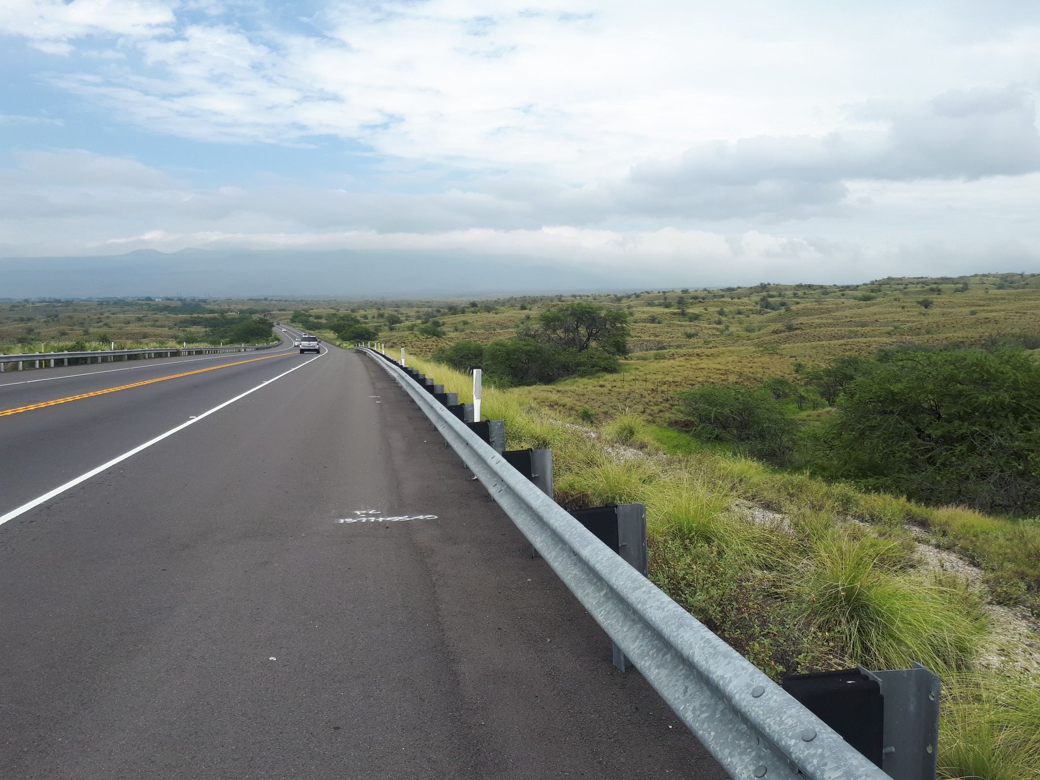 A car drives on a highway through arid fields under a cloudy sky. - Lulahala Point to Waimea loop