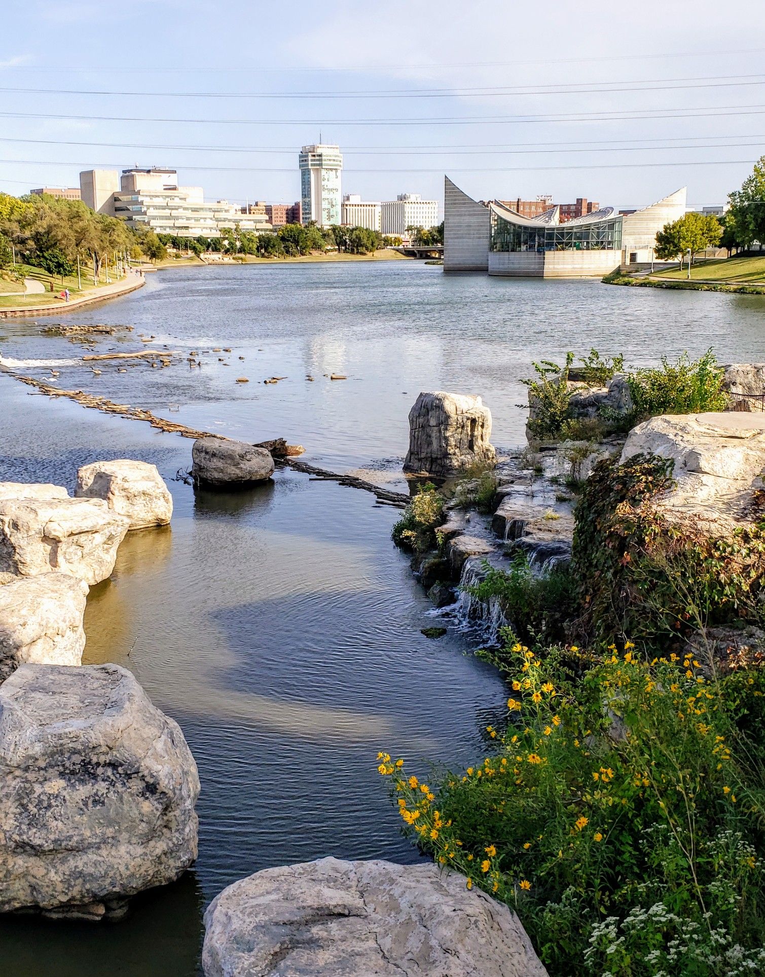 Urban river flows past rocky banks with distant city buildings under bright sky. - Great Plains Nature Center – Keeper of the Plains Statue loop from Colonel James Jabara Airport