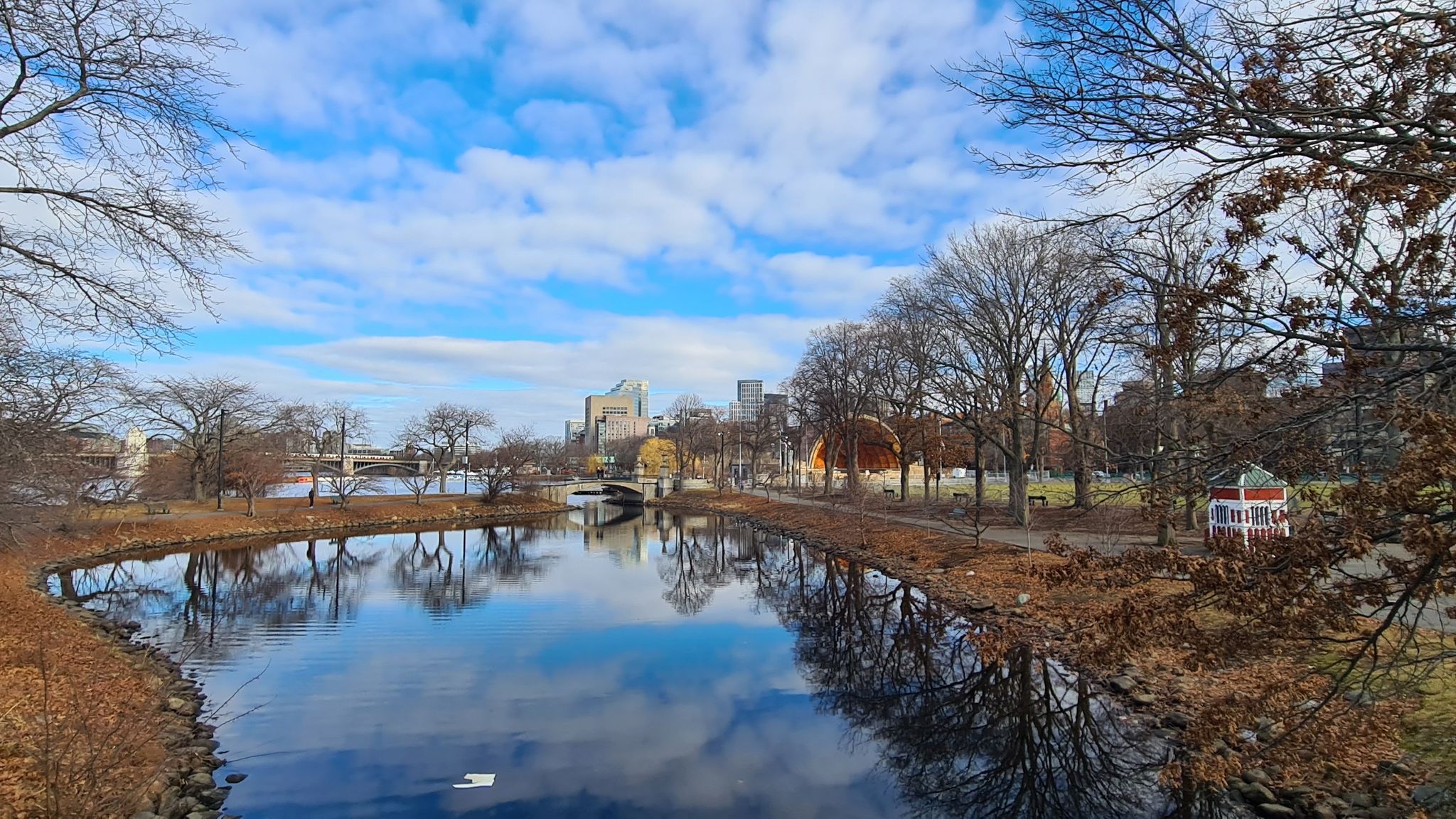 River reflecting a cloudy sky, with bare trees and distant city buildings. - Storrow Lagoon – Southern Promenade on Charles River loop from Boston University Central