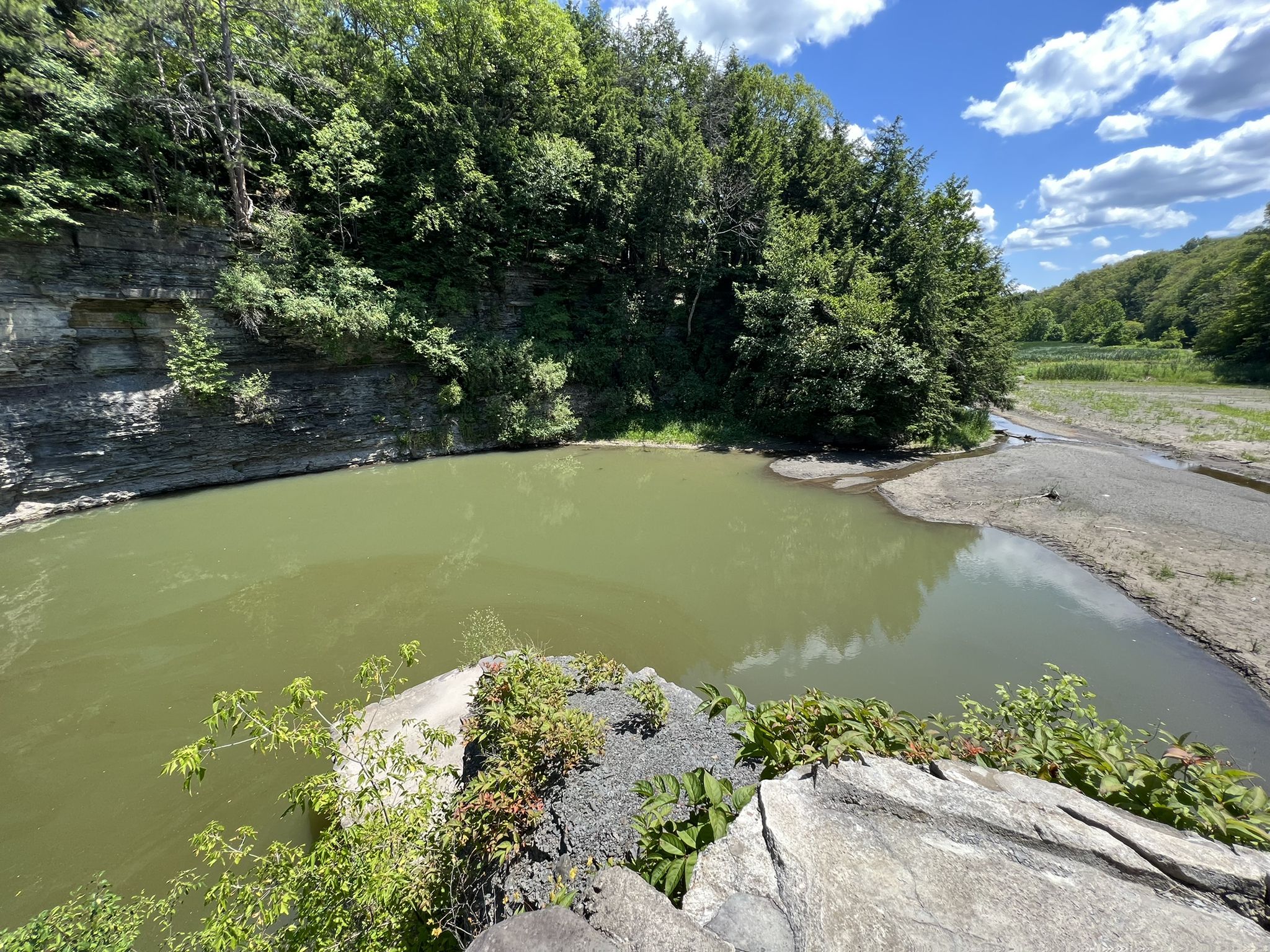 Green river pool beside a rocky cliff and sandy bank, sunny day. - Lake Treman Loop