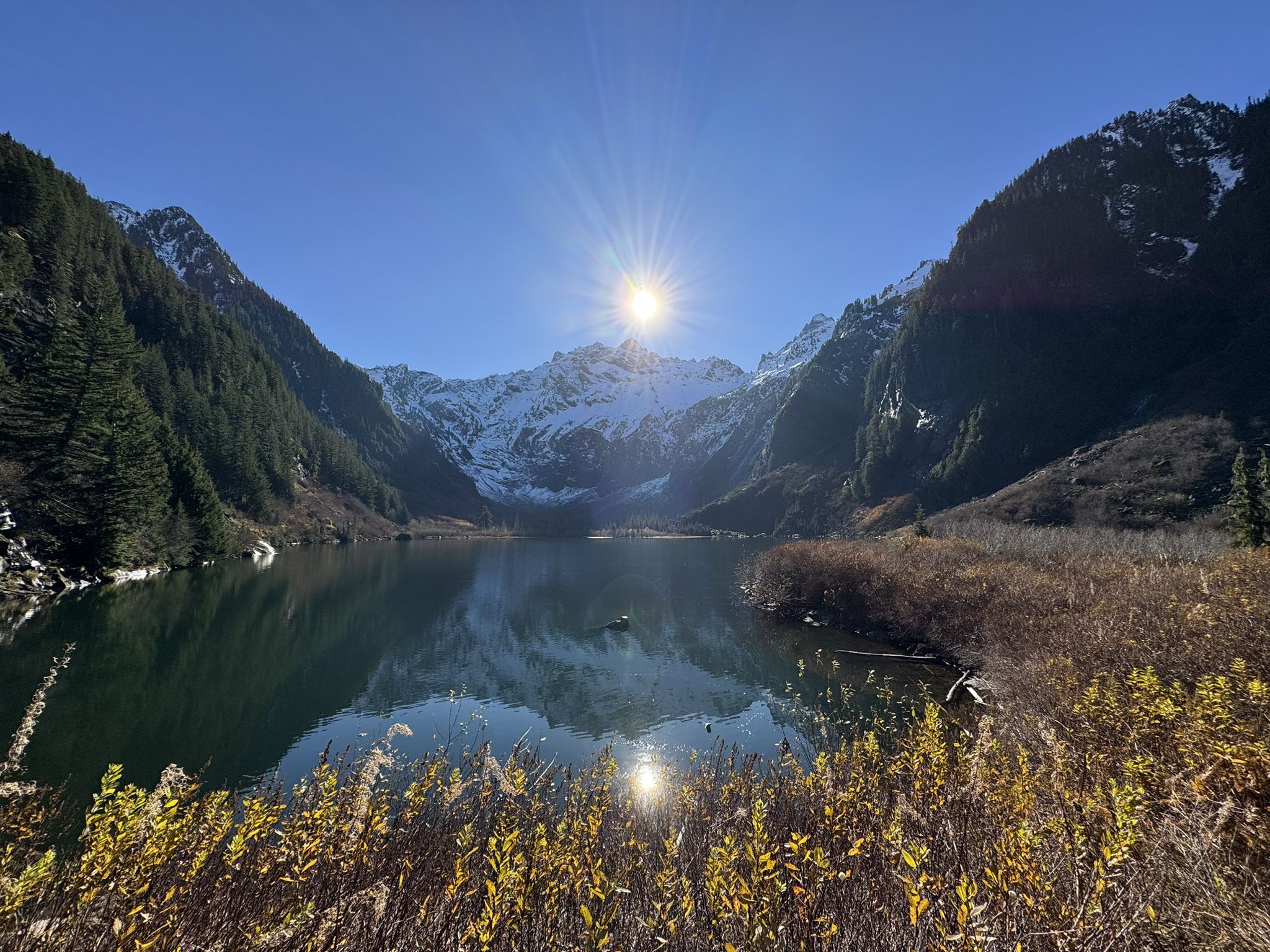 Bright sunburst over a mountain lake reflecting snowy peaks and autumn plants. - Elliot Creek (Goat Lake) Trail