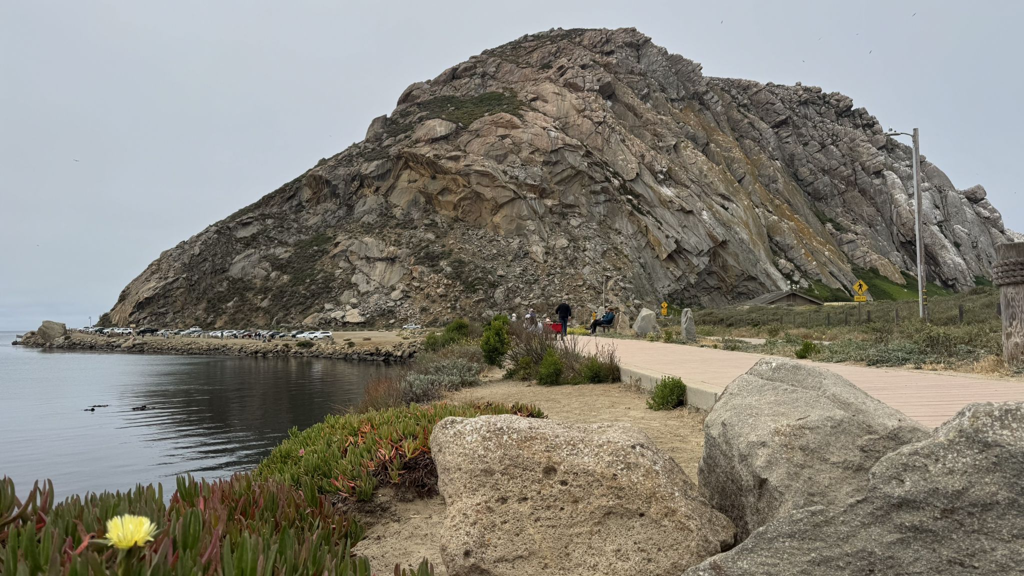 Large rocky hill beside calm water with people on a boardwalk. - Cloisters to Morro Rock Trail