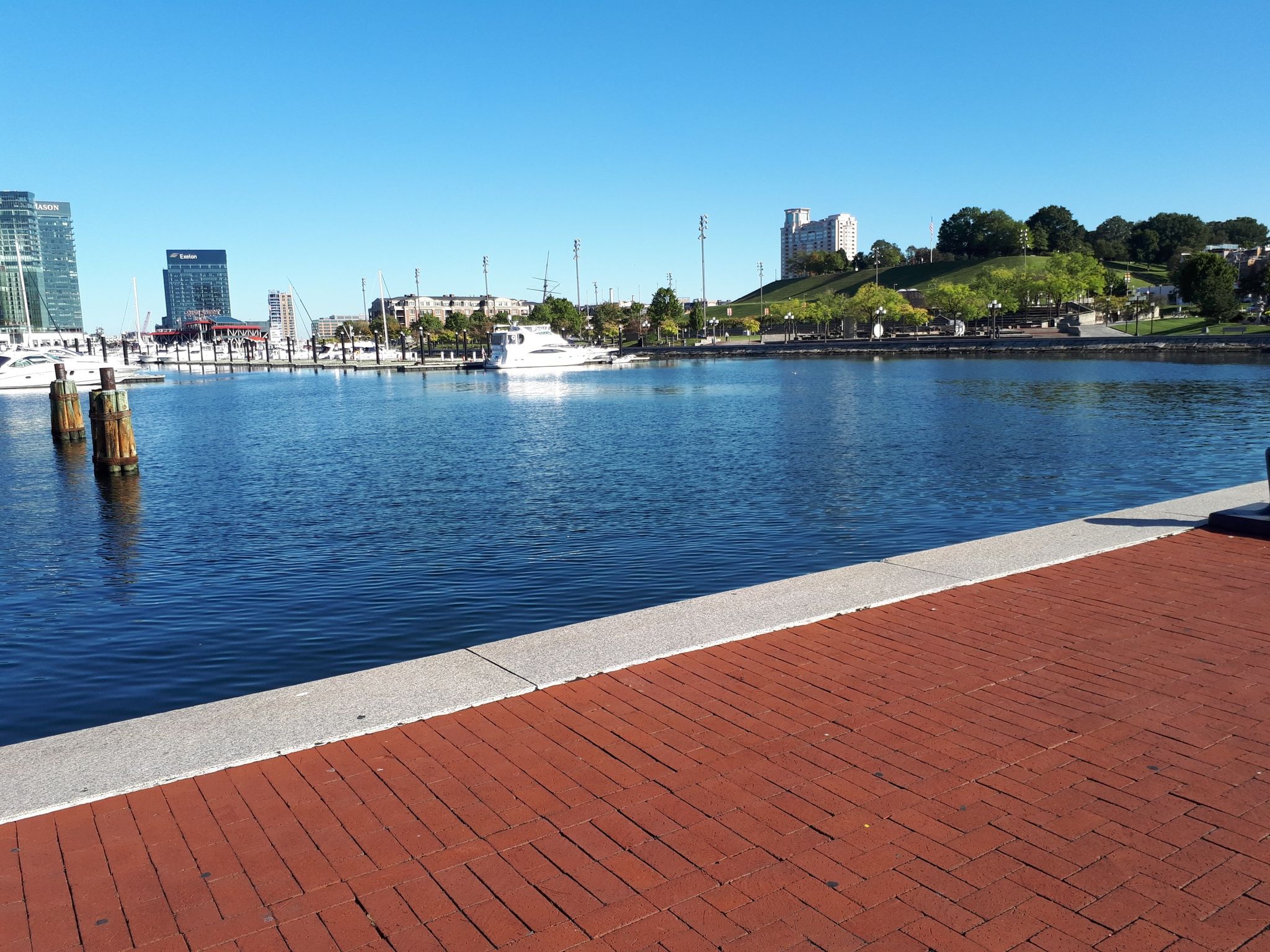 A brick promenade by calm water, with distant city buildings and boats under blue sky. - Baltimore Waterfront – Baltimore Waterfront Promenade loop from Baltimore