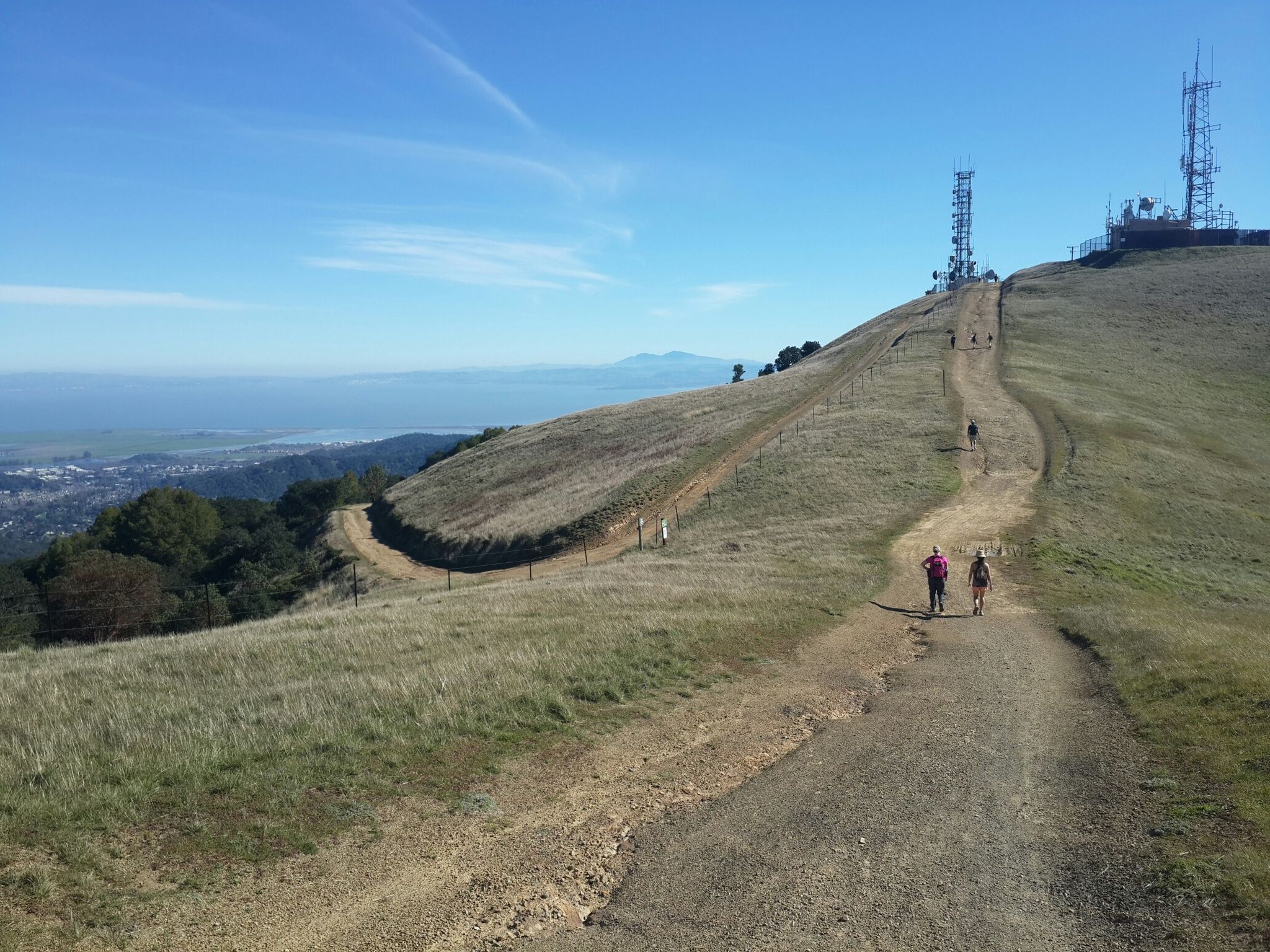 Two people walk on a dirt path up a grassy hill towards communication towers. - Big Rock Ridge Trail