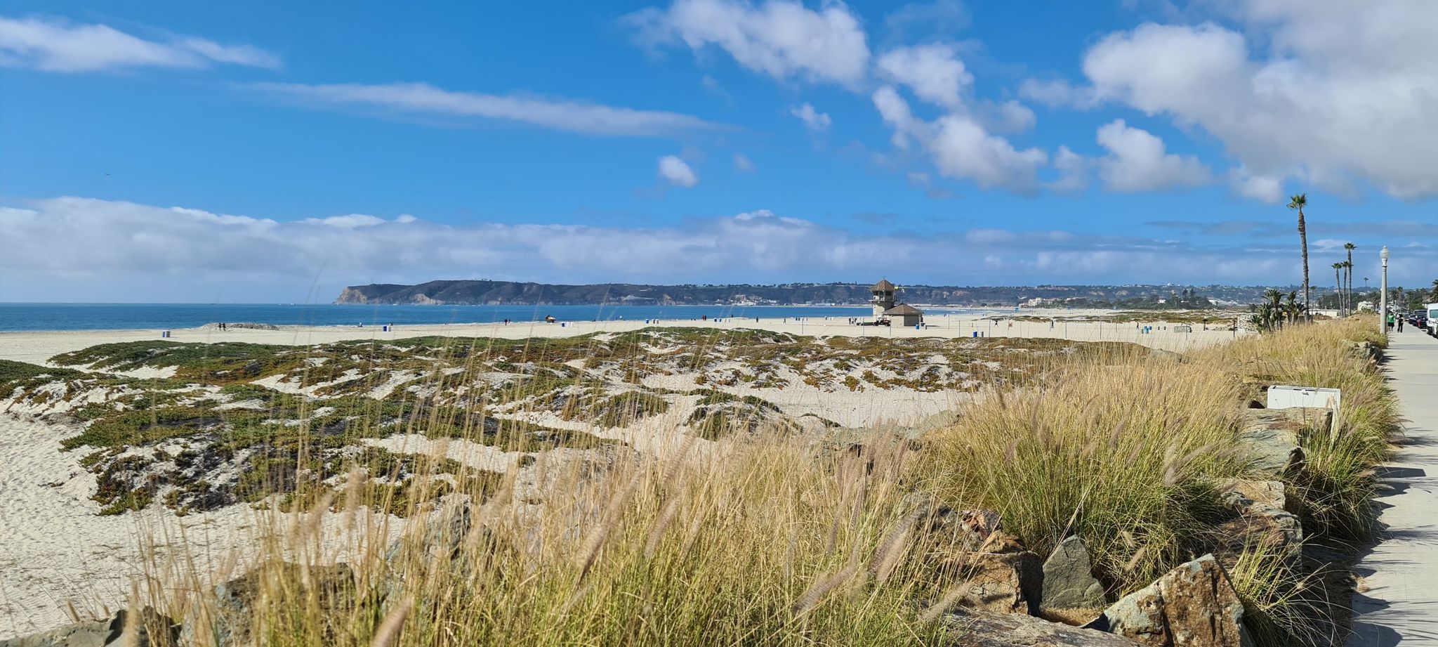 Wide sandy beach with dunes, ocean, and distant land under a bright, cloudy sky. - Coronado Beach