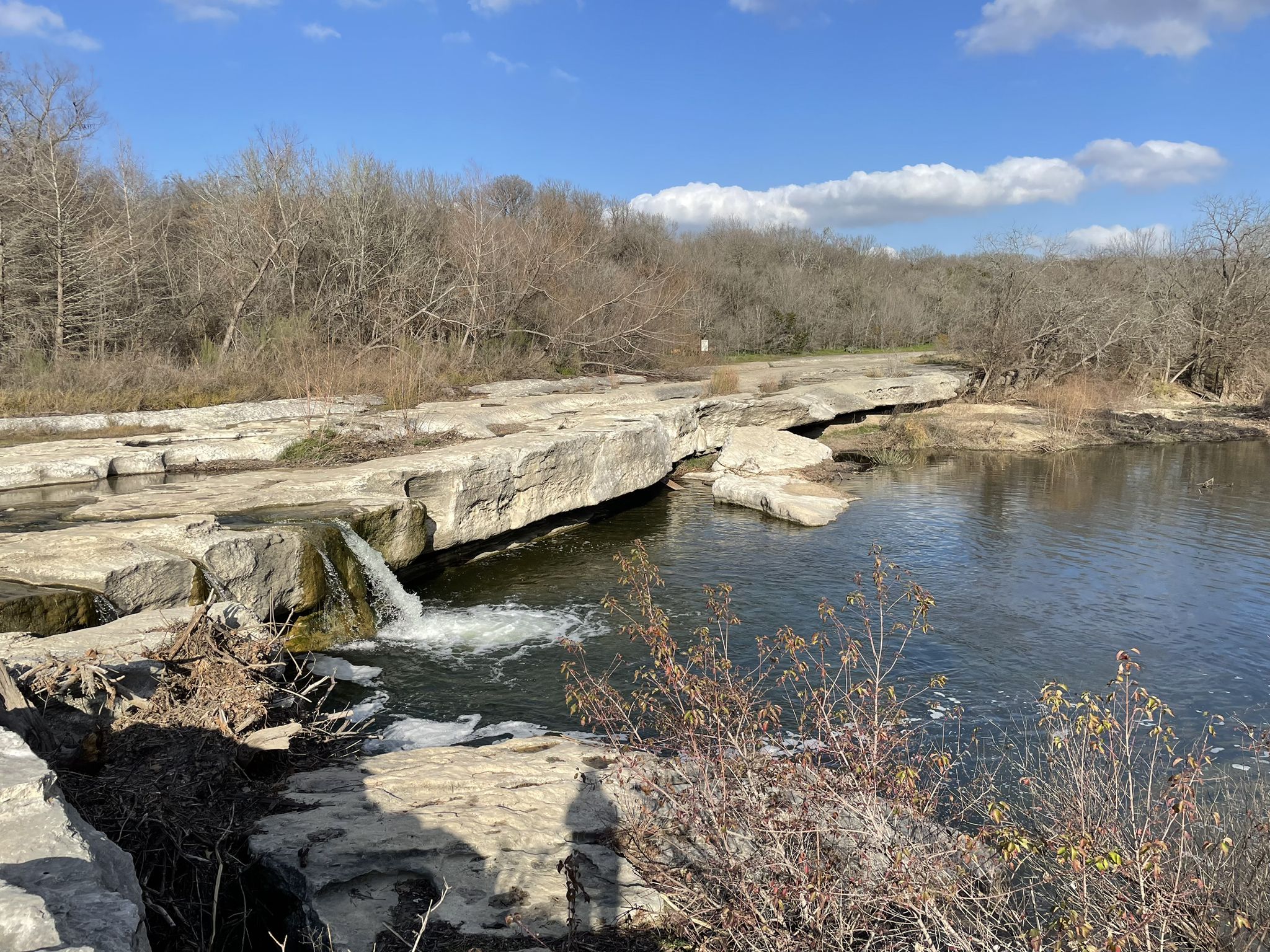 Small waterfall over rocky ledges into a river, bare trees, sunny day. - Homestead Trail