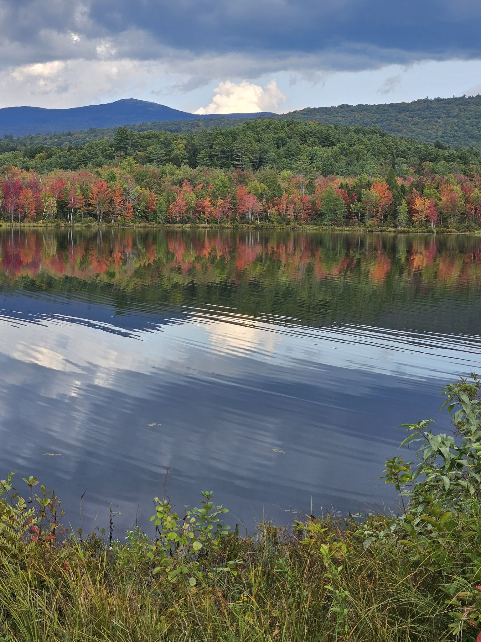 Lake reflecting vibrant autumn trees and distant mountains under a cloudy sky. - Mud Pond loop from Canaan
