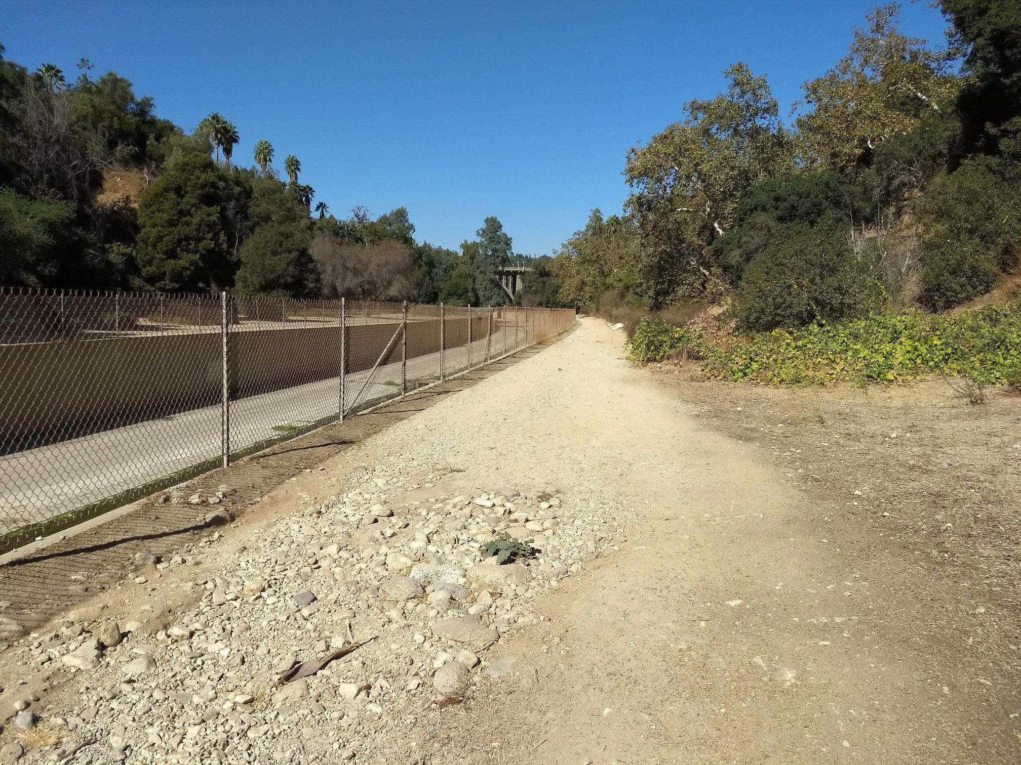 A sunny dirt path runs alongside a chain-link fence through a dry, wooded landscape. - Lower Arroyo Seco Trail