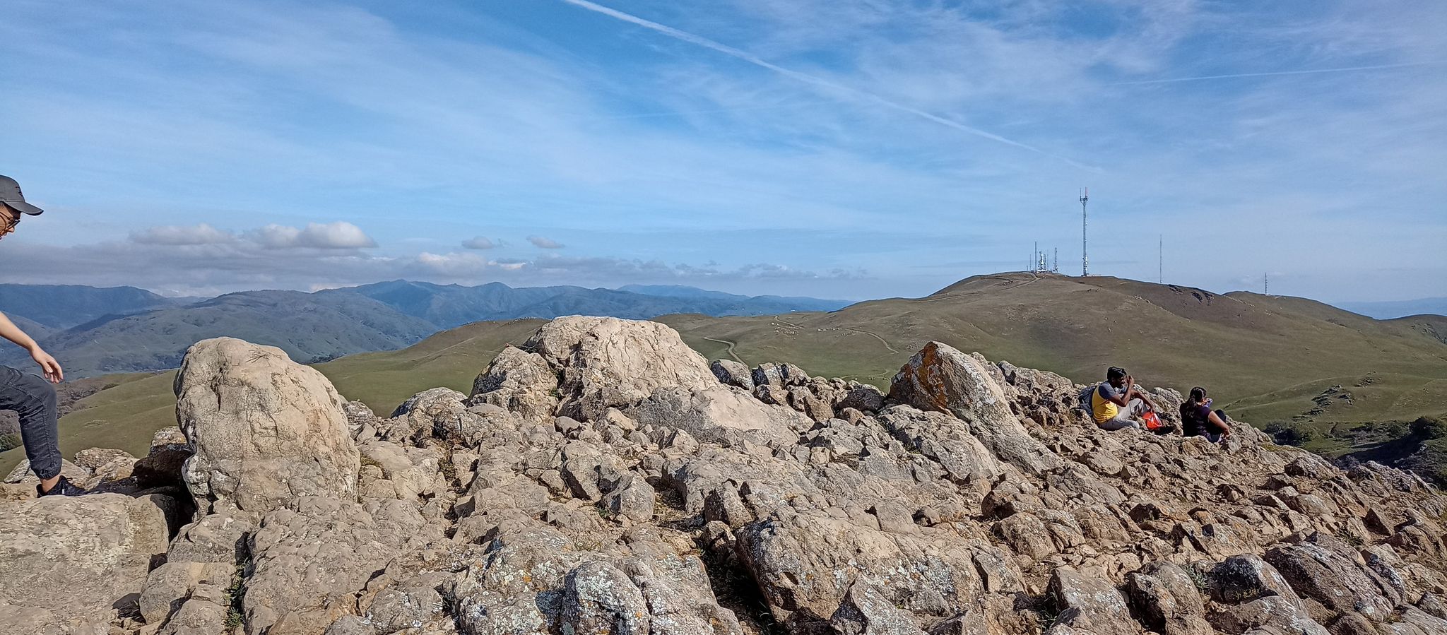 People rest on a rocky mountain ridge with distant green hills and towers. - Mission Peak via Ohlone Wilderness Trail