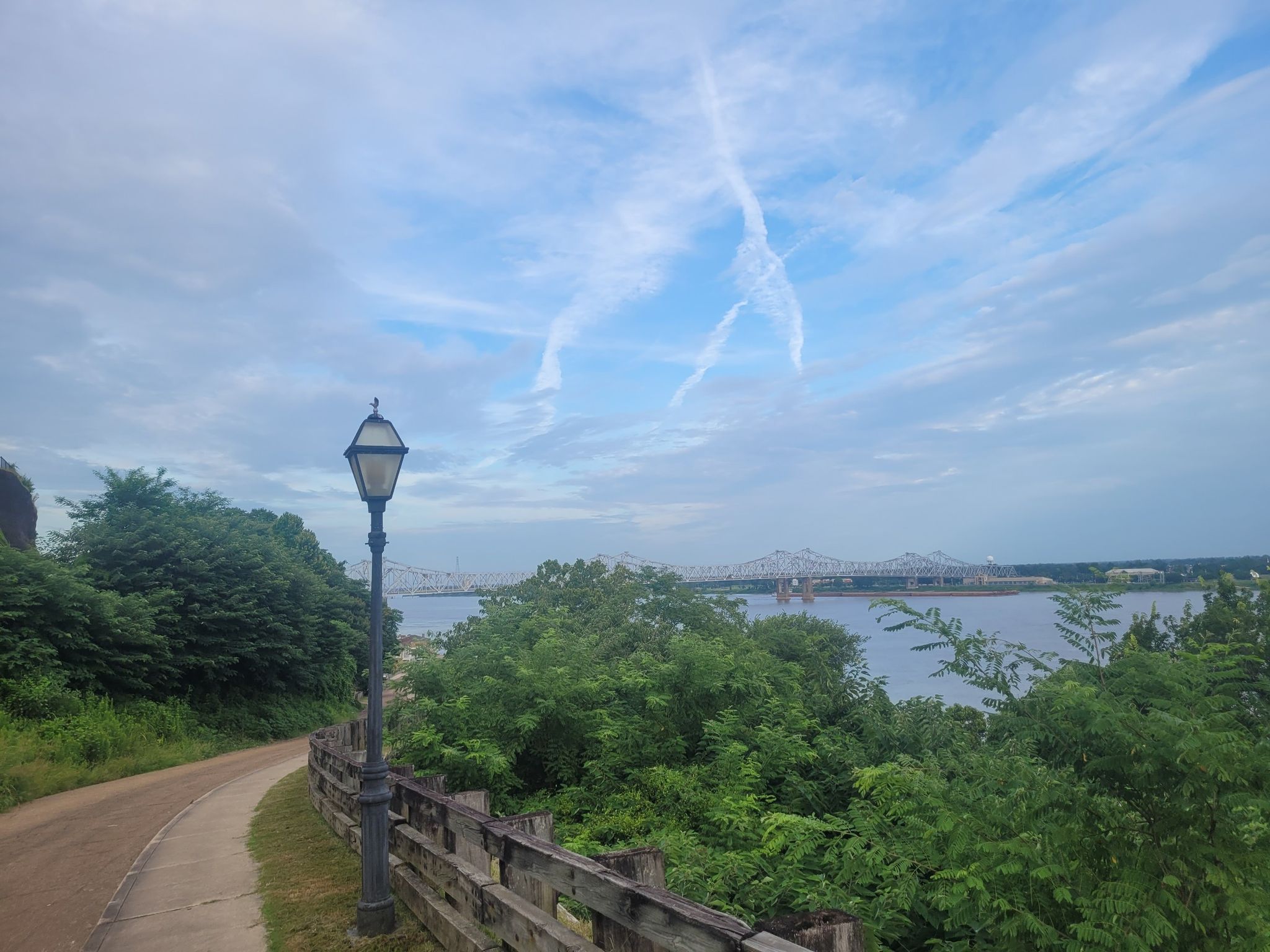 Paved path, lamppost, green trees, river, and distant bridge under a blue sky. - Pontura Historic House – Stanton Hall loop from Natchez