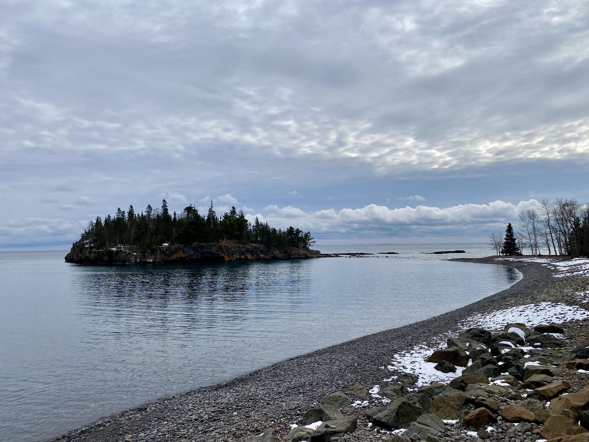Cloudy day over a rocky beach with snow, calm water, and a forested island. - Split Rock Lighthouse – View of Split Rock Lighthouse loop from Split Rock Lighthouse
