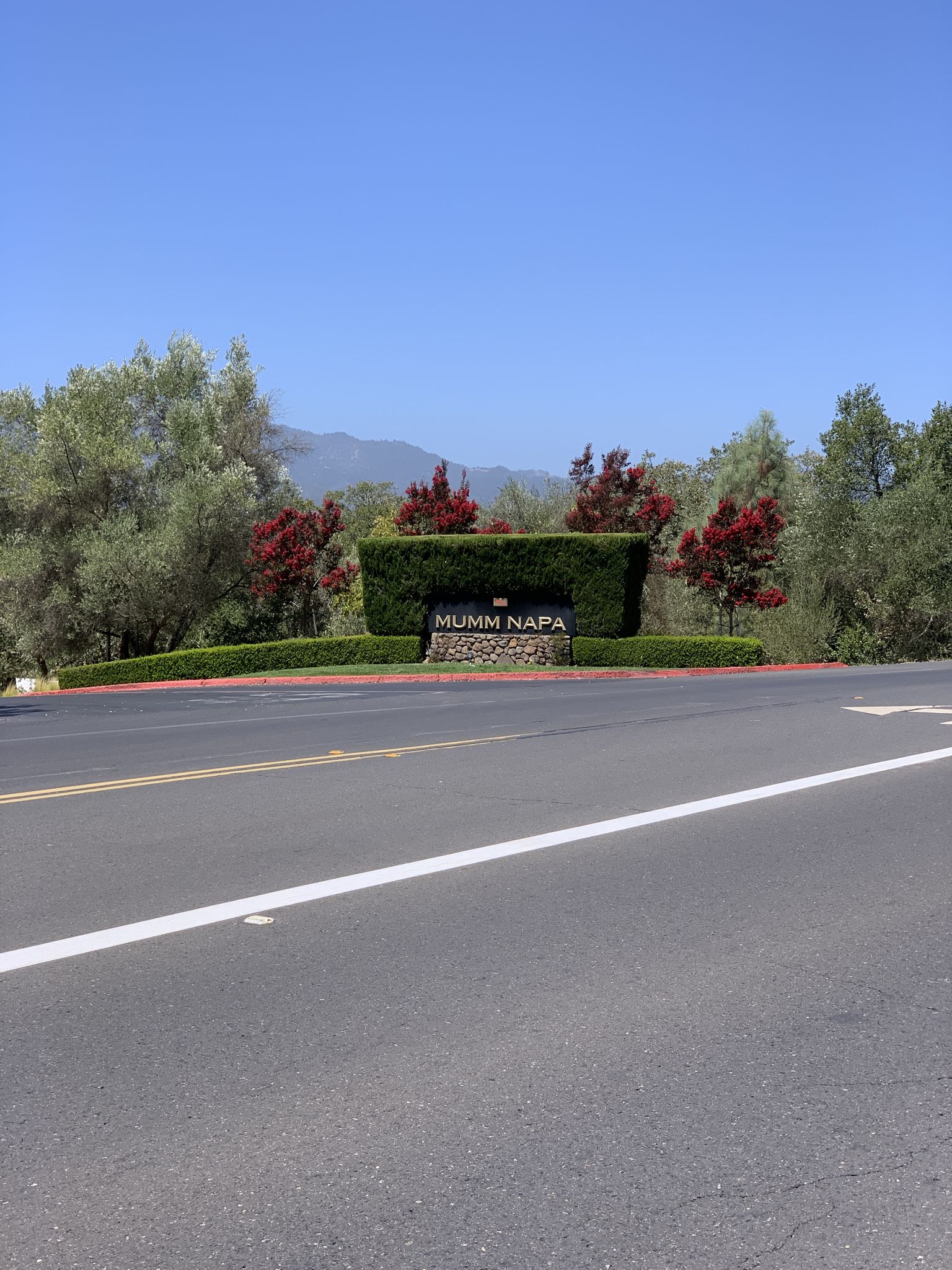 Paved road leading to a landscaped entrance with a sign, trees, and bushes. - Saint Helena – Welcome to Napa Valley Sign loop from Saint Helena