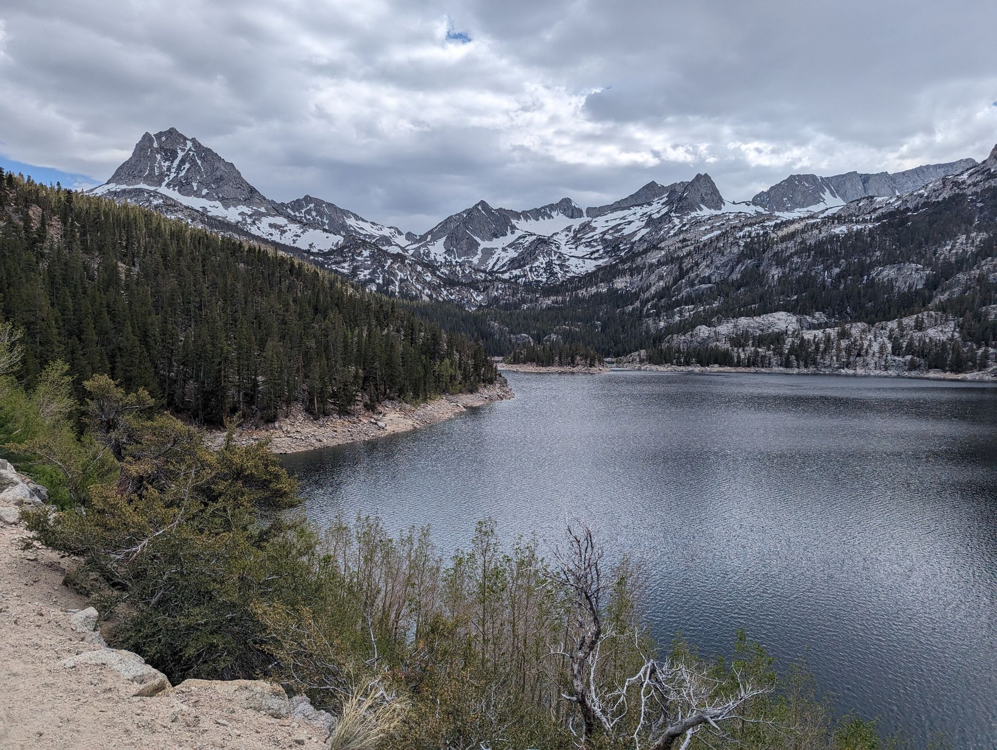 A large lake surrounded by pine forests and snow-capped mountains under a cloudy sky. - Bull Lake via Bishop Pass Trail