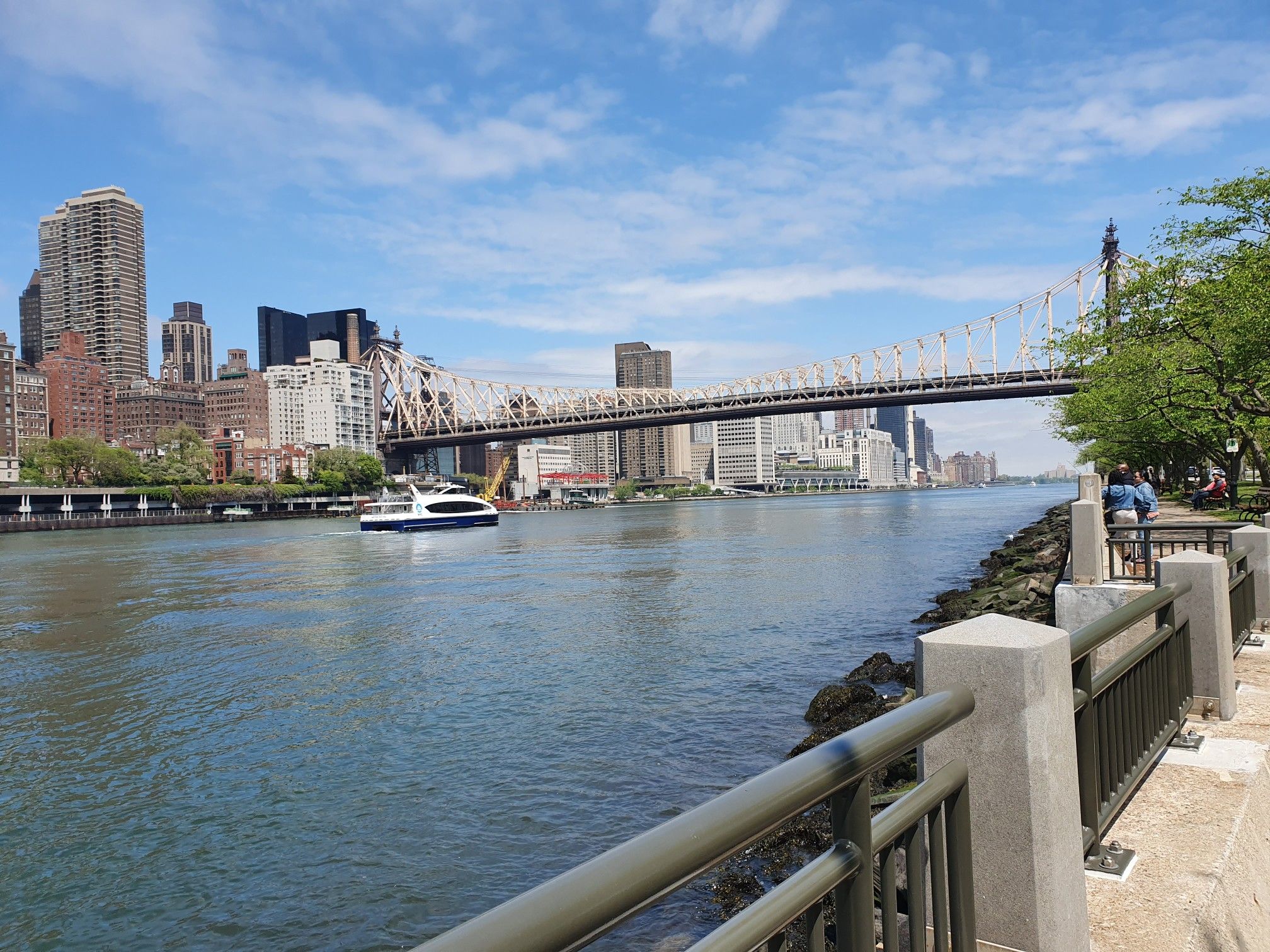Large bridge over a river with a boat, city skyline, and person on path. - Roosevelt Island Tramway – Southpoint Park loop from Queens Plaza