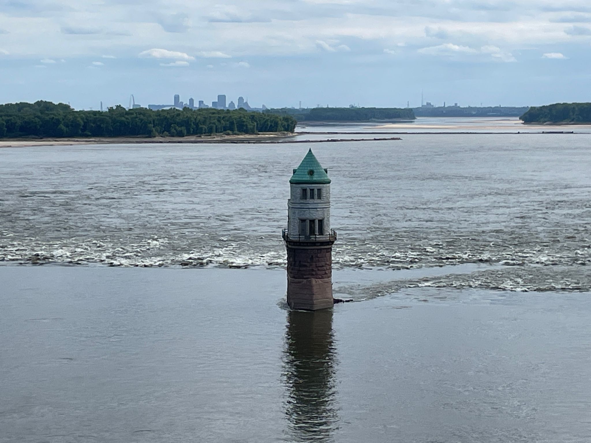 A stone lighthouse stands in choppy river water with a distant city. - Old Chain of Rocks Bridge – Old Chain of Rocks Bridge loop from Granite City