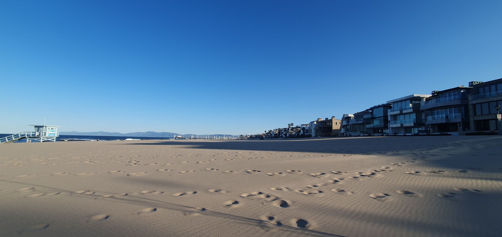 Wide sandy beach with footprints, ocean, and houses under a clear blue sky. - Noble Park loop from Manhattan Beach