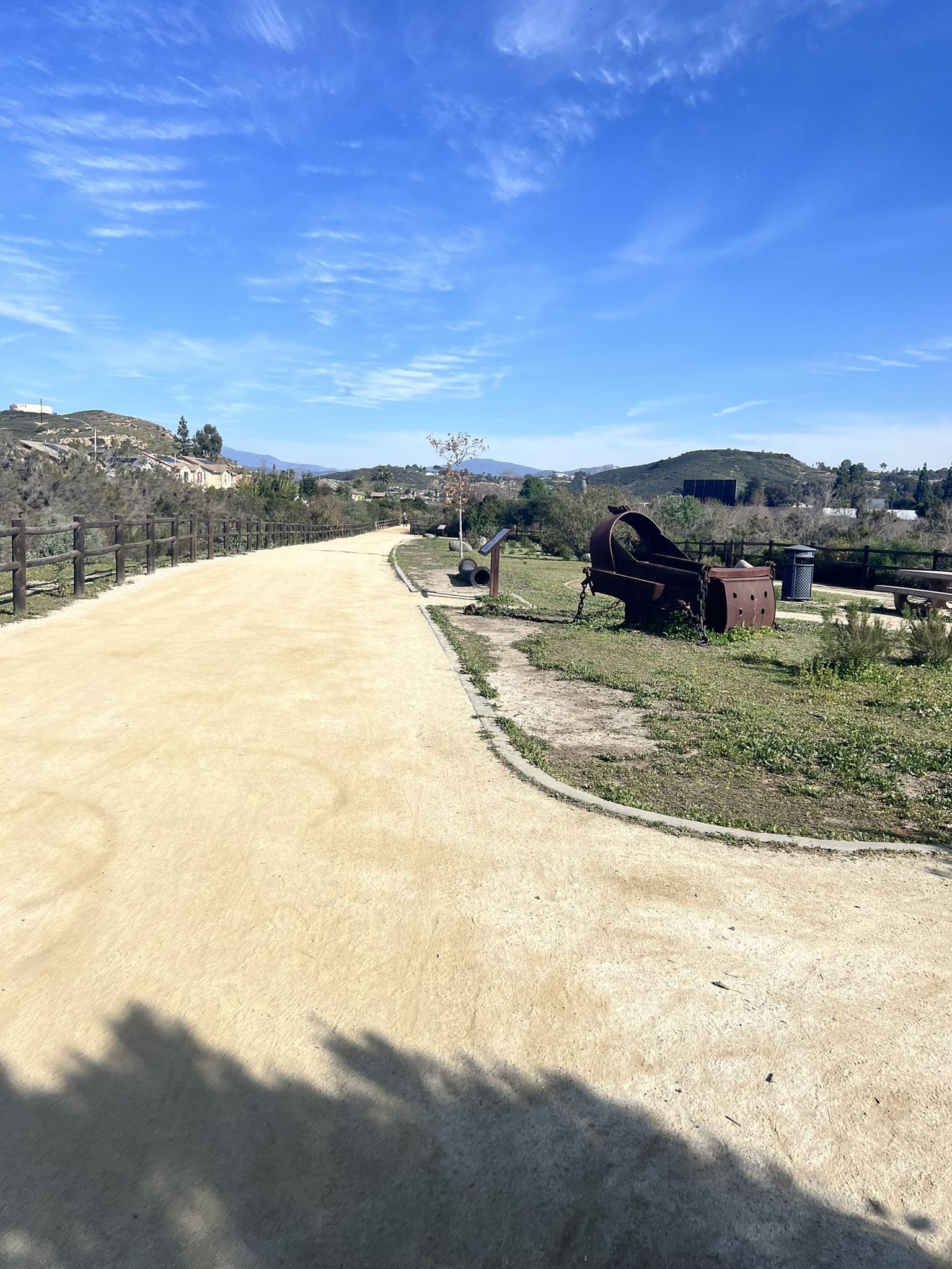 Wide, light-colored path curving right, with hills, rusty art, and blue sky. - San Diego River Trail
