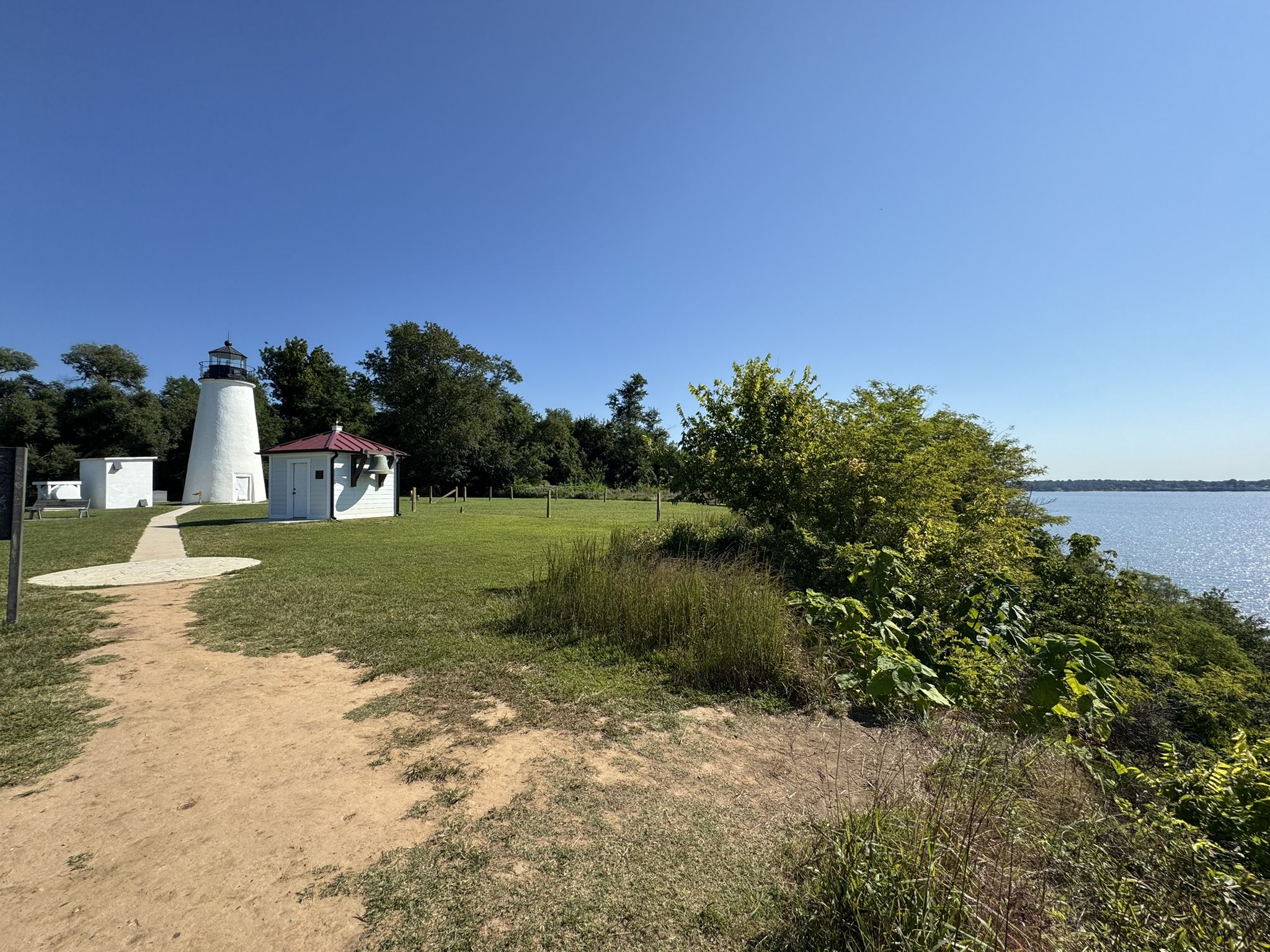 White lighthouse and buildings on a grassy bank by water under a clear sky. - Turkey Point Lighthouse Trail