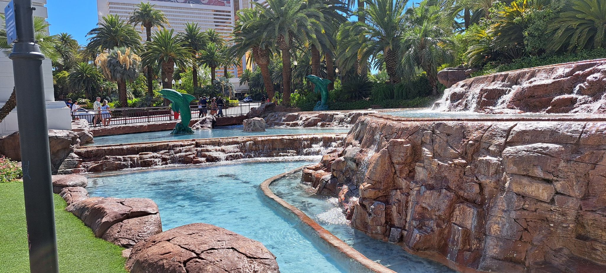 Multi-tiered artificial waterfall and pools with palm trees and buildings. - The MGM Grand – The Venetian Resort loop from Las Vegas Valley