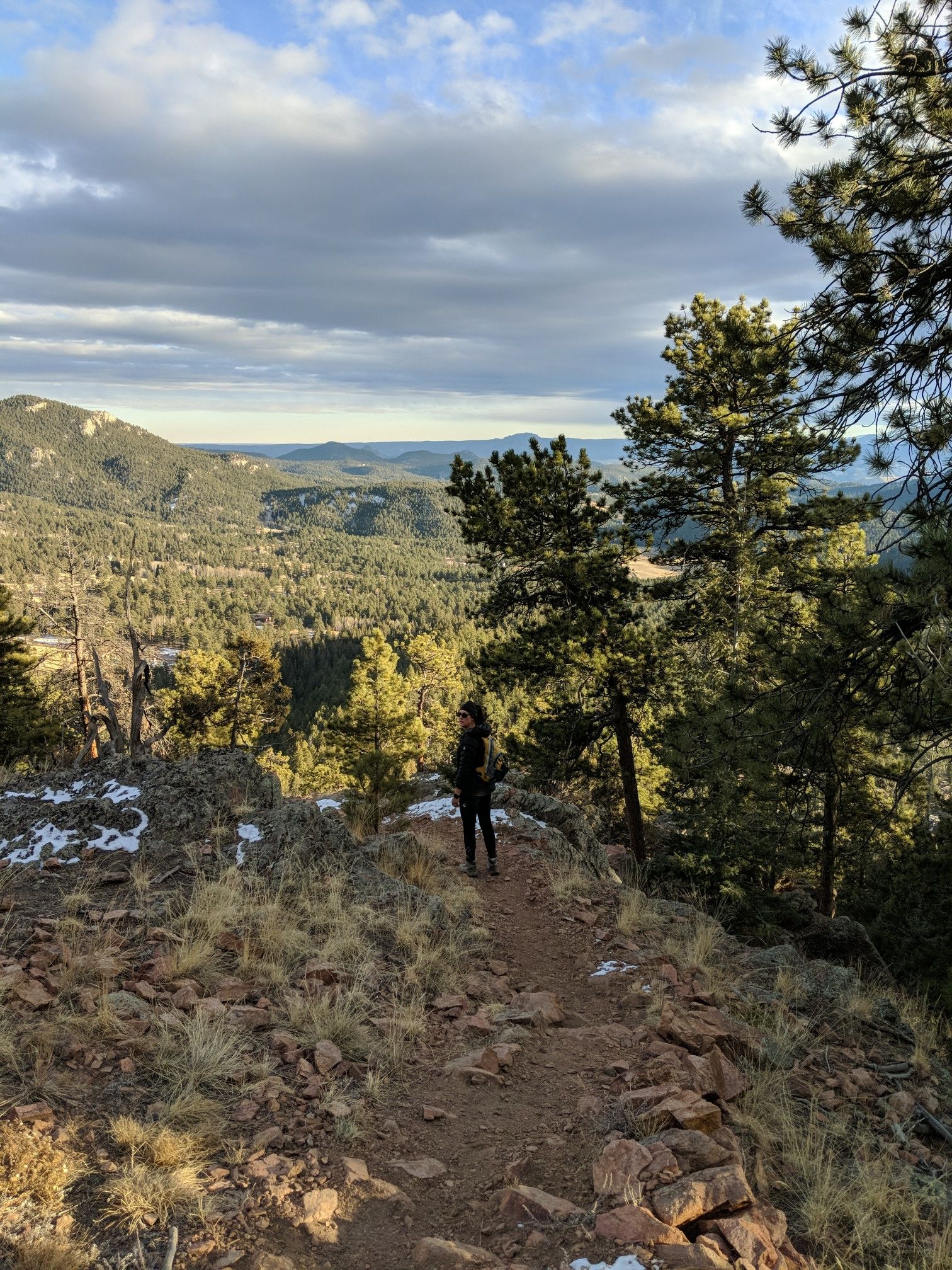 A person stands on a rocky trail overlooking a vast mountain landscape. - Scoutline Trail loop from Lazy V