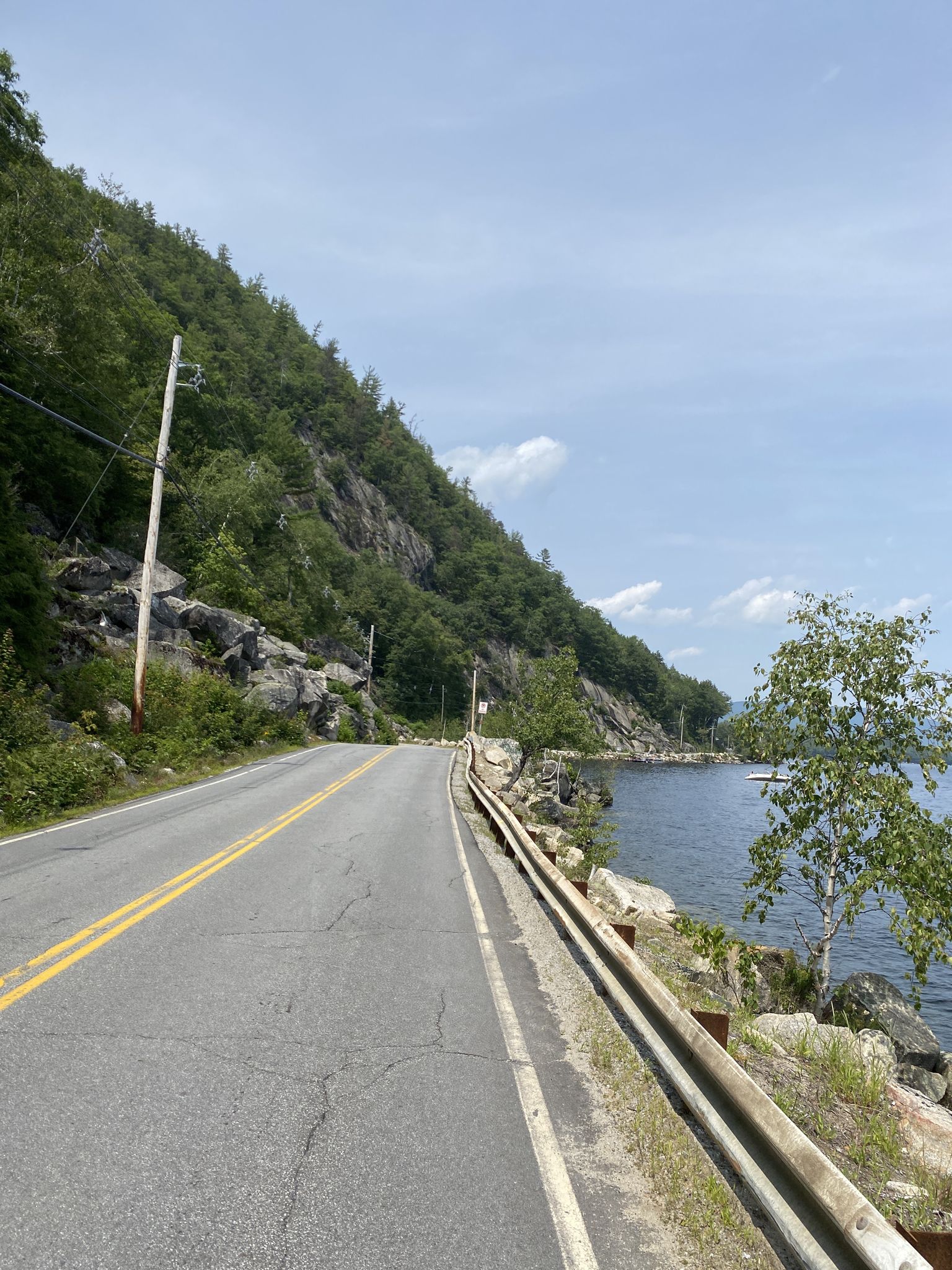 Road alongside a steep, rocky, forested mountain next to a lake. - West Shore Road – Wellington Beach State Park loop from Common Man