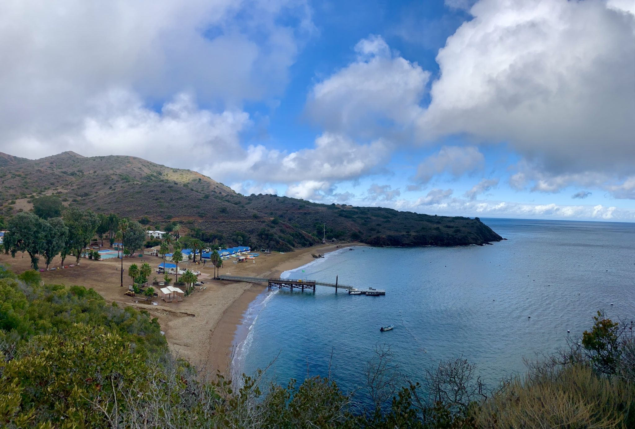 Coastal bay with a pier, sandy beach, and buildings under a bright sky. - View of Big Geiger Cove – View of Cherry Cove loop from Indian Rock