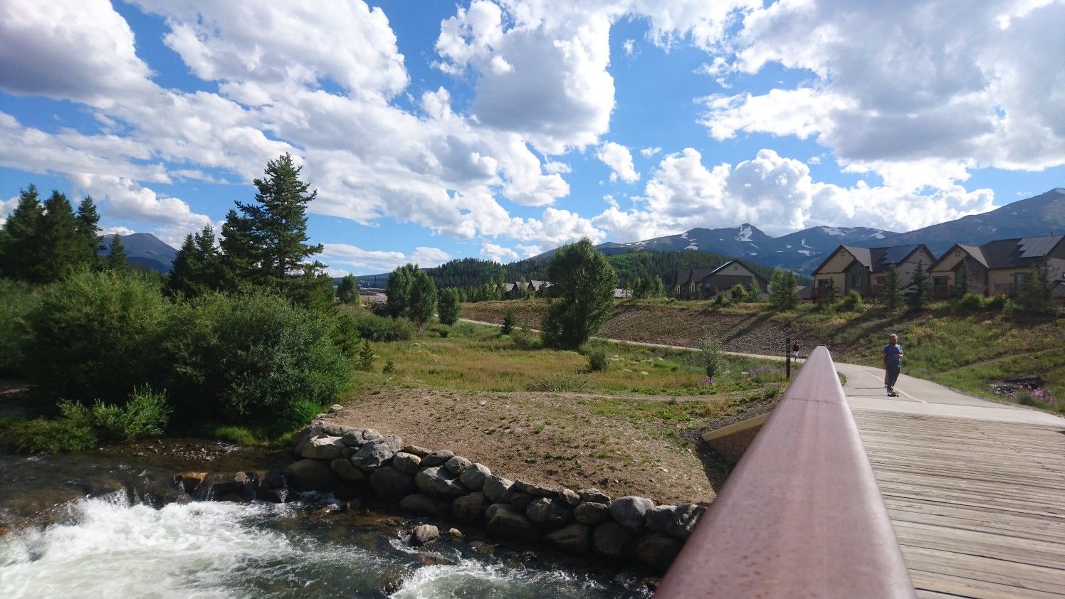 A person stands on a wooden bridge over a rushing river, with houses and mountains. - Blue River Rec Path