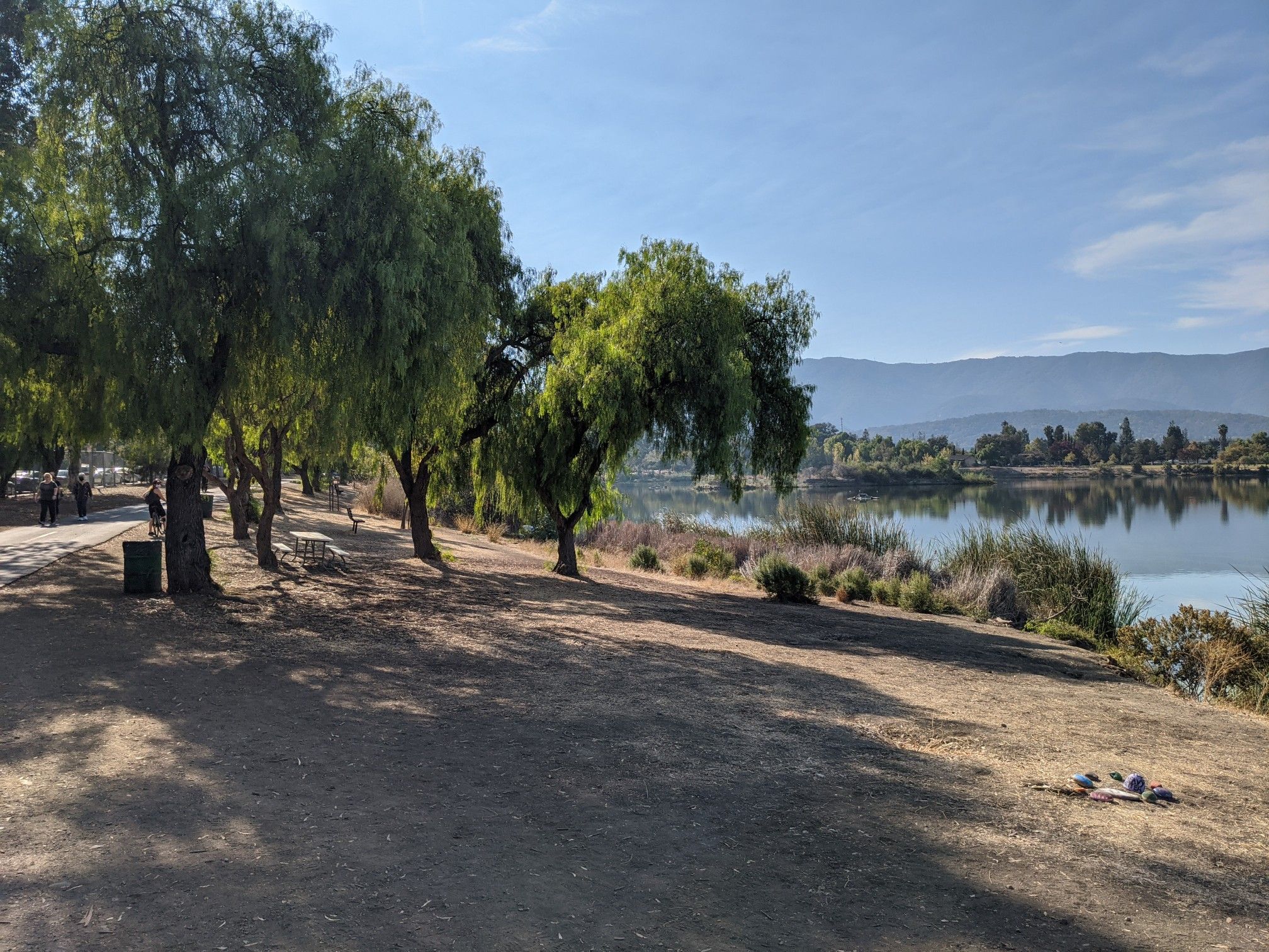 Sunny lakeside path with trees, distant mountains, and people walking. - Pfeiffer Park – Almaden Lake Trail loop from Almaden Valley