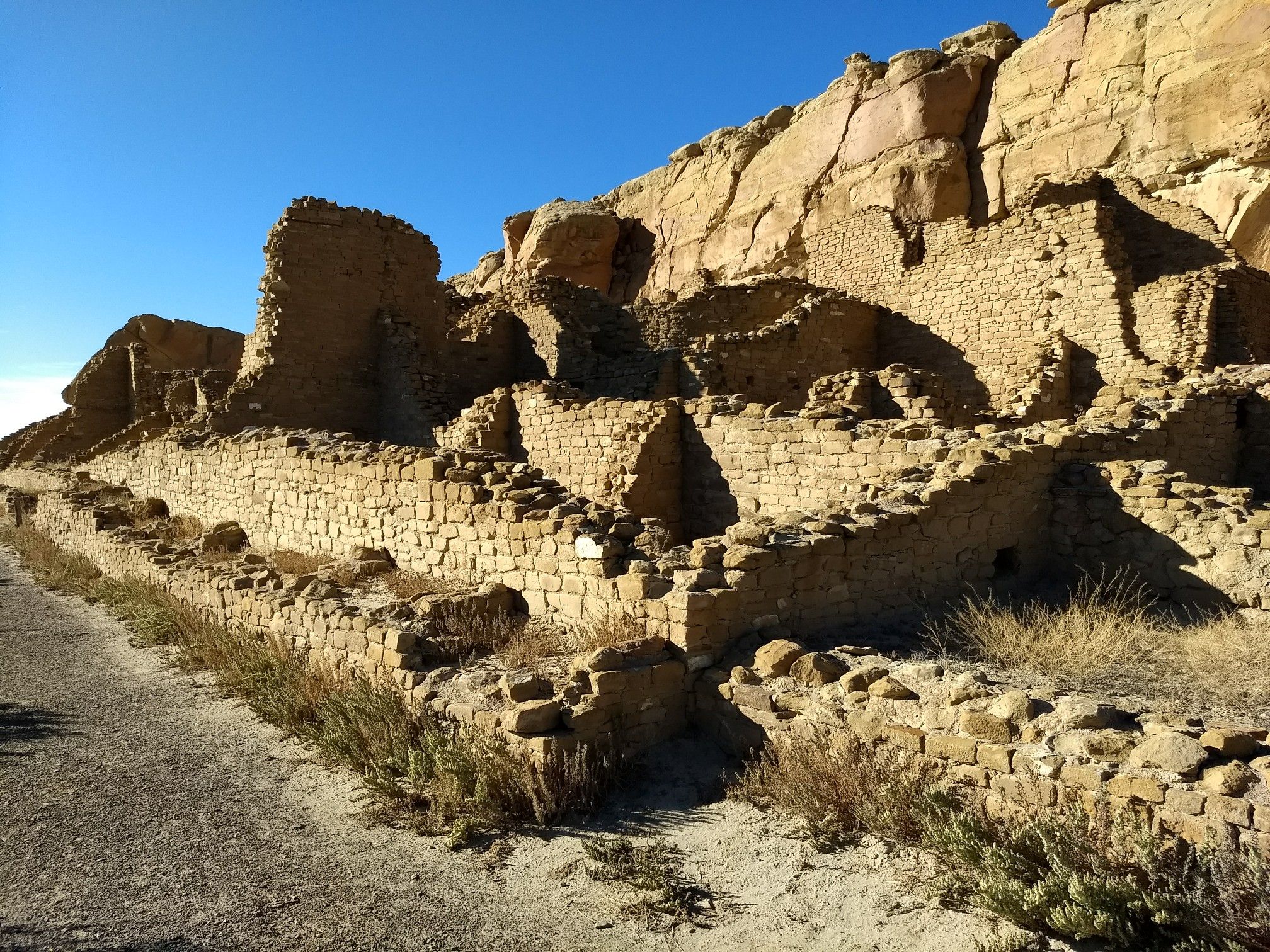 Ancient stone walls built into a rocky cliff face under bright sunlight. - Kin Kletso – Penasco Blanco loop from Pueblo Bonito Great House