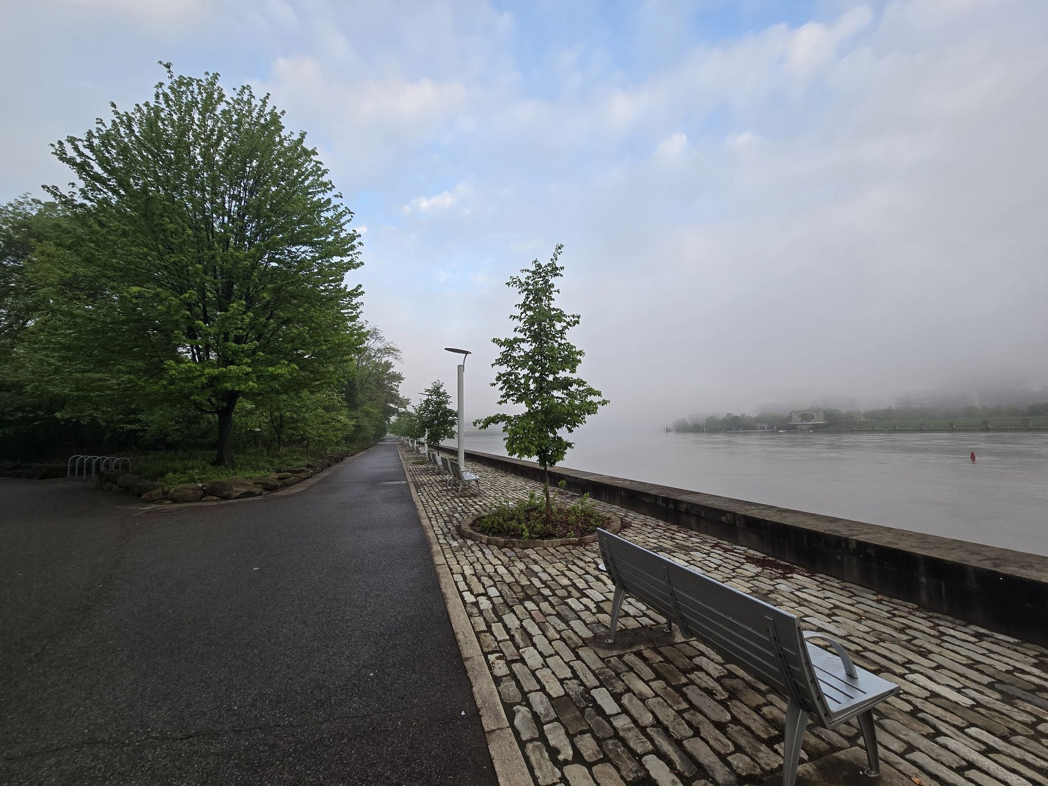 Cobblestone path and benches beside a foggy river, green trees. - Fort Pitt Block House – Point State Park Fountain loop from Pittsburgh
